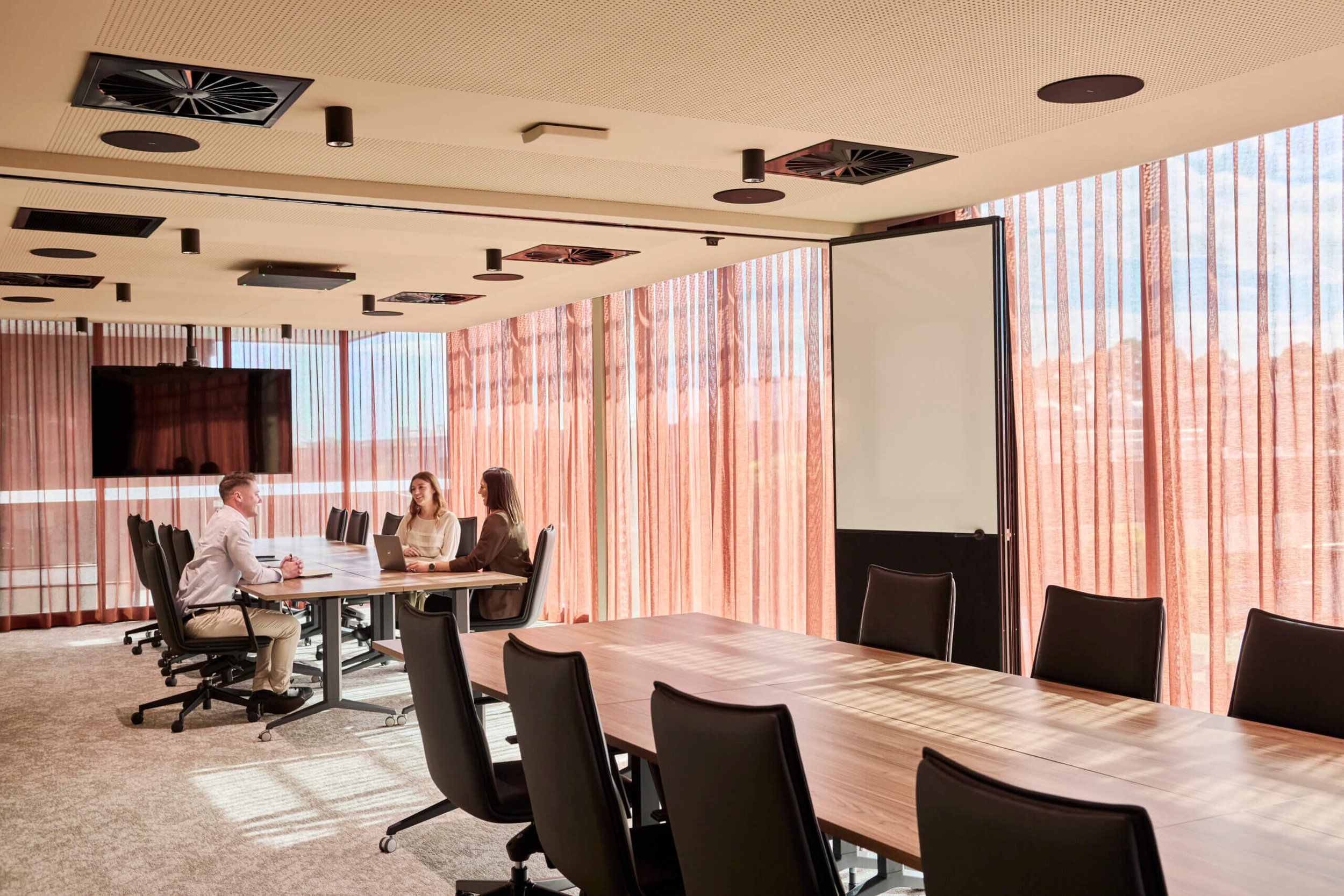 A conference room with a long wooden table, black chairs, a wall-mounted TV, large windows with pink curtains, and three people sitting at the table having a discussion.
