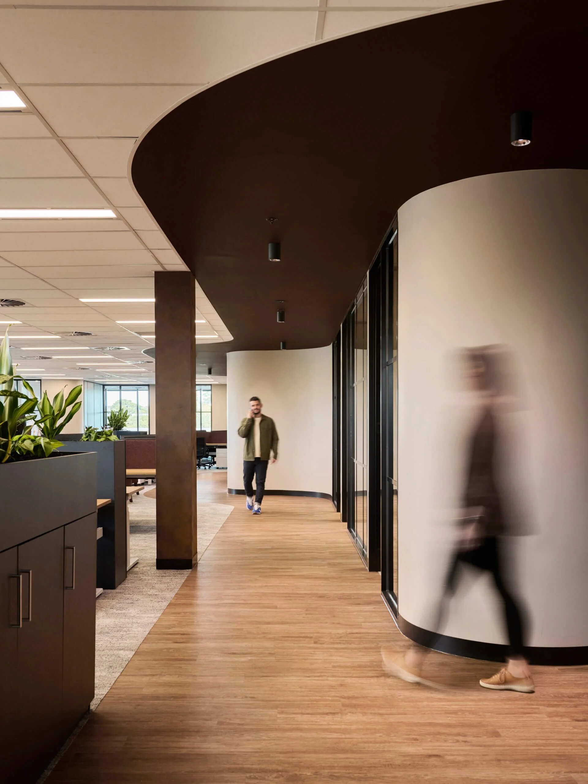 Modern office interior with a bar-height wood floor, a curved gray wall, black-framed glass doors, and plants. Two people are walking through, one in the foreground with blurred motion, and another in the background talking on their phone.