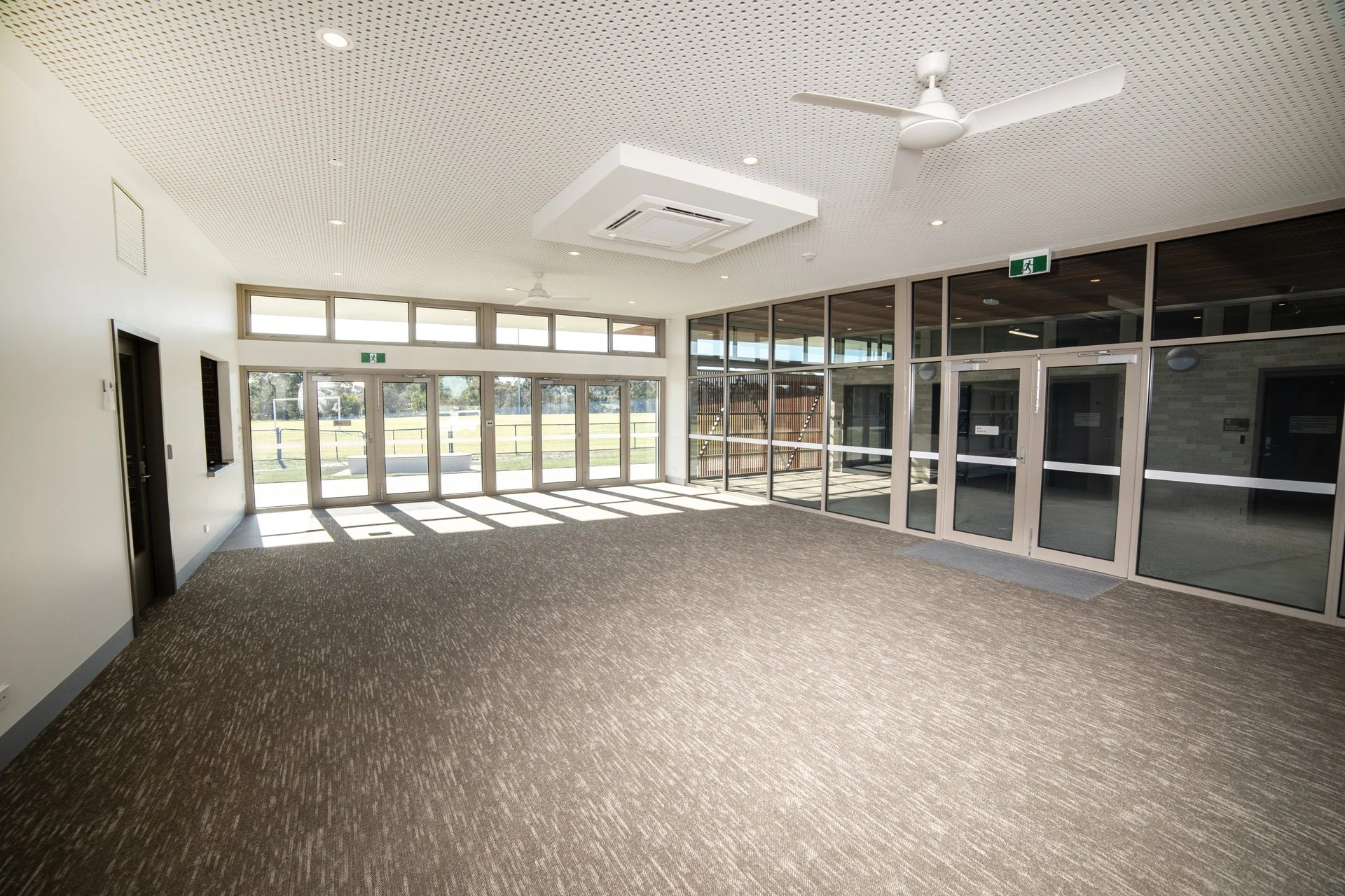 Empty indoor space with glass doors and windows, ceiling fans, and an emergency exit sign, overlooking a field outside.