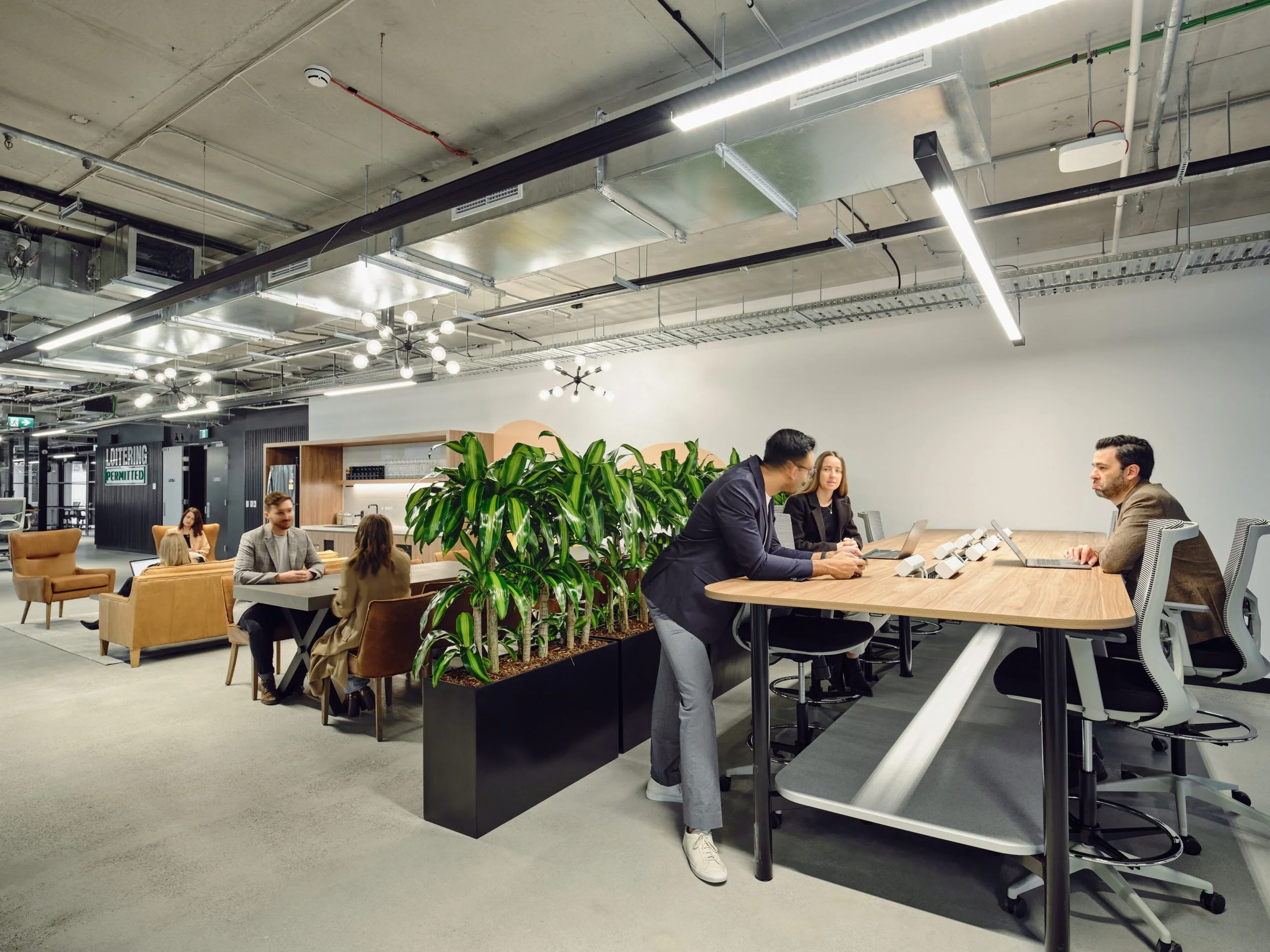 An office or coworking space with people working and talking at a large wooden table. The room features modern lighting, plants, and an industrial ceiling with exposed ducts and pipes.