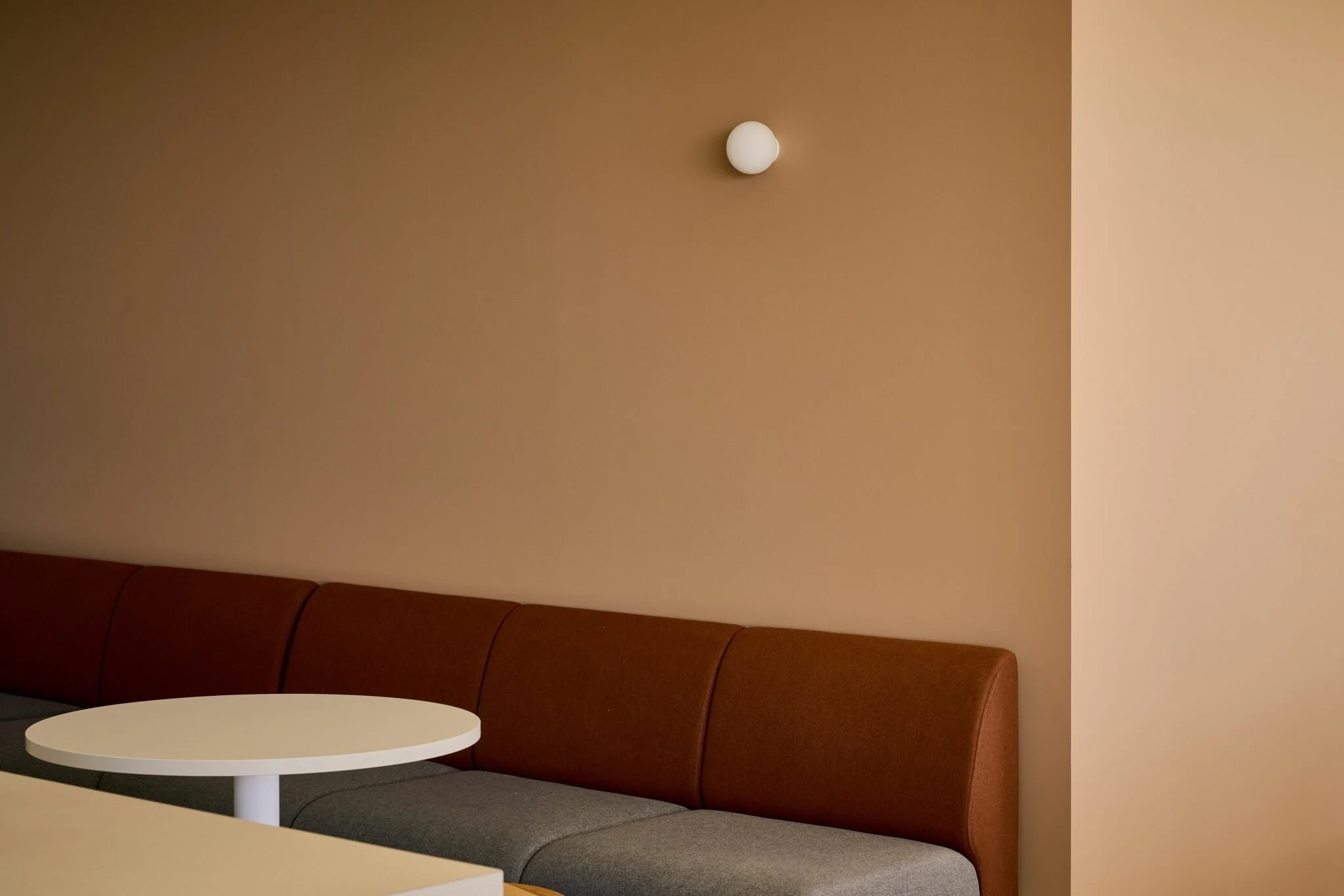 A corner of a room with a brown upholstered bench, a white oval table, beige walls, and a small wall-mounted light fixture.