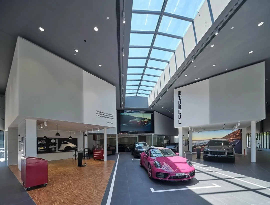 Modern Porsche dealership showroom with a pink sports car in the foreground, and several other cars, large screens, and Porsche branding visible.