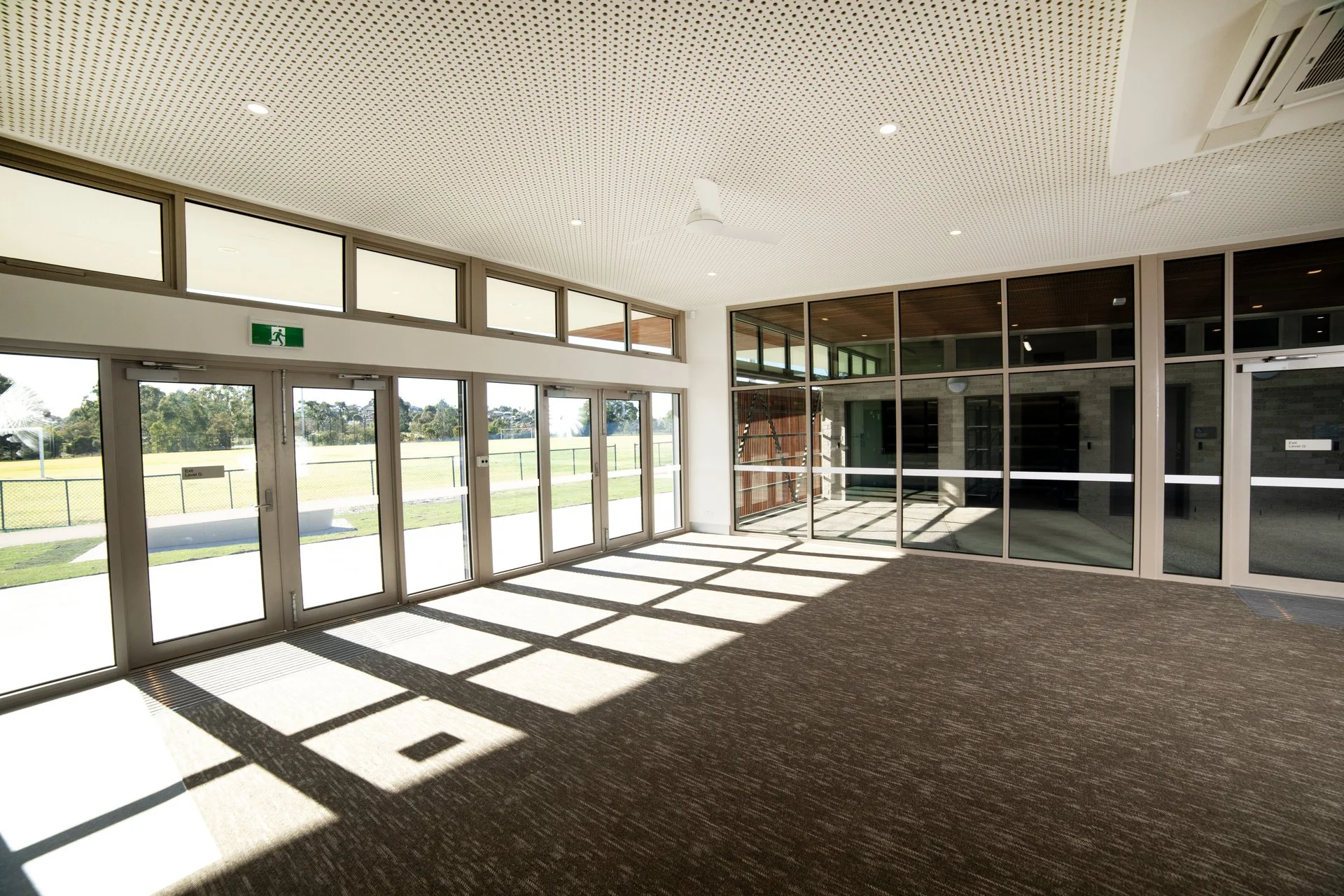 Interior of a building with large glass doors and windows, views of a grassy sports field outside, featuring an exit sign above and a ceiling fan.