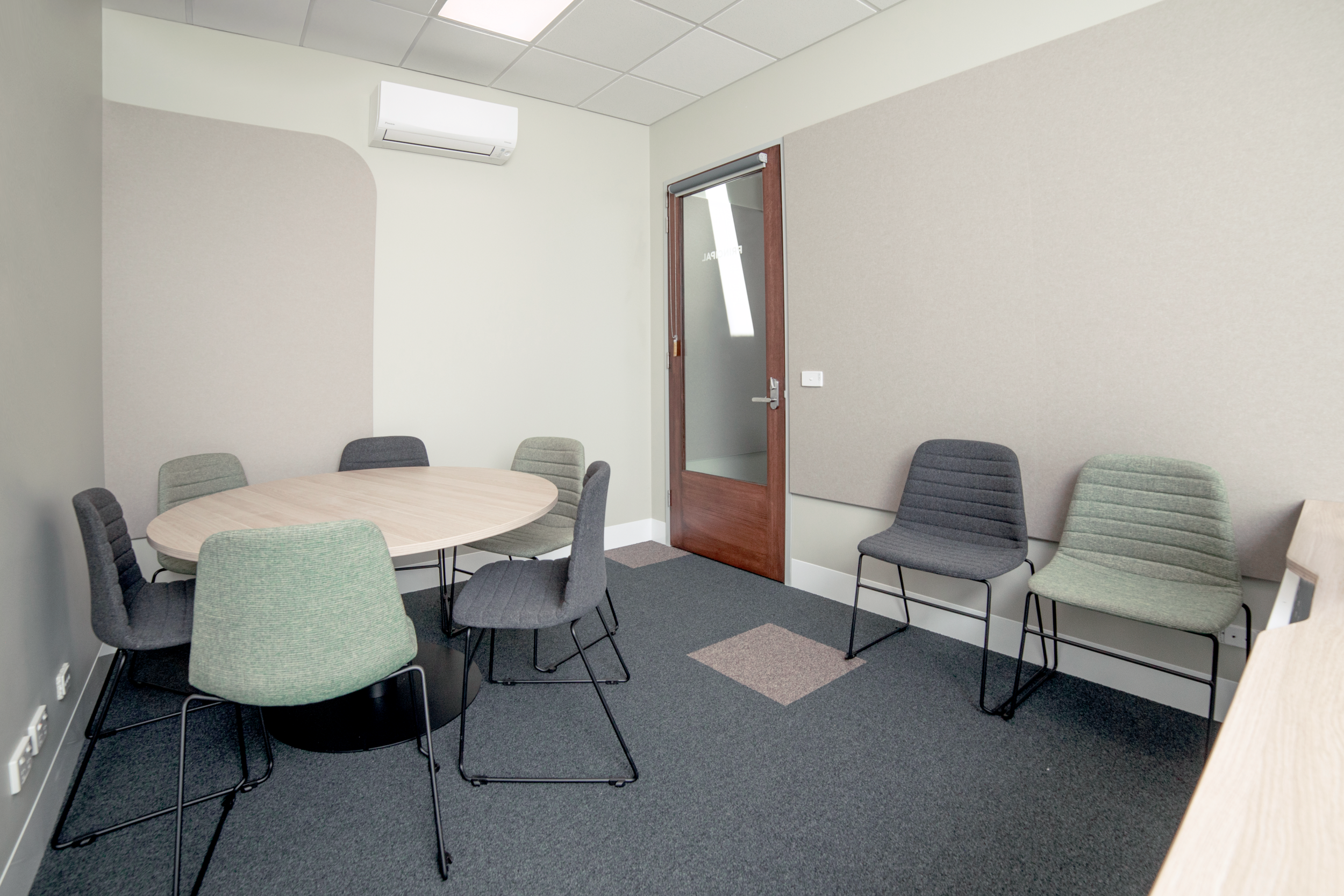 An empty conference or meeting room with a round table surrounded by eight chairs, a wooden door with a glass window, a wall-mounted air conditioner, and gray wall panels.