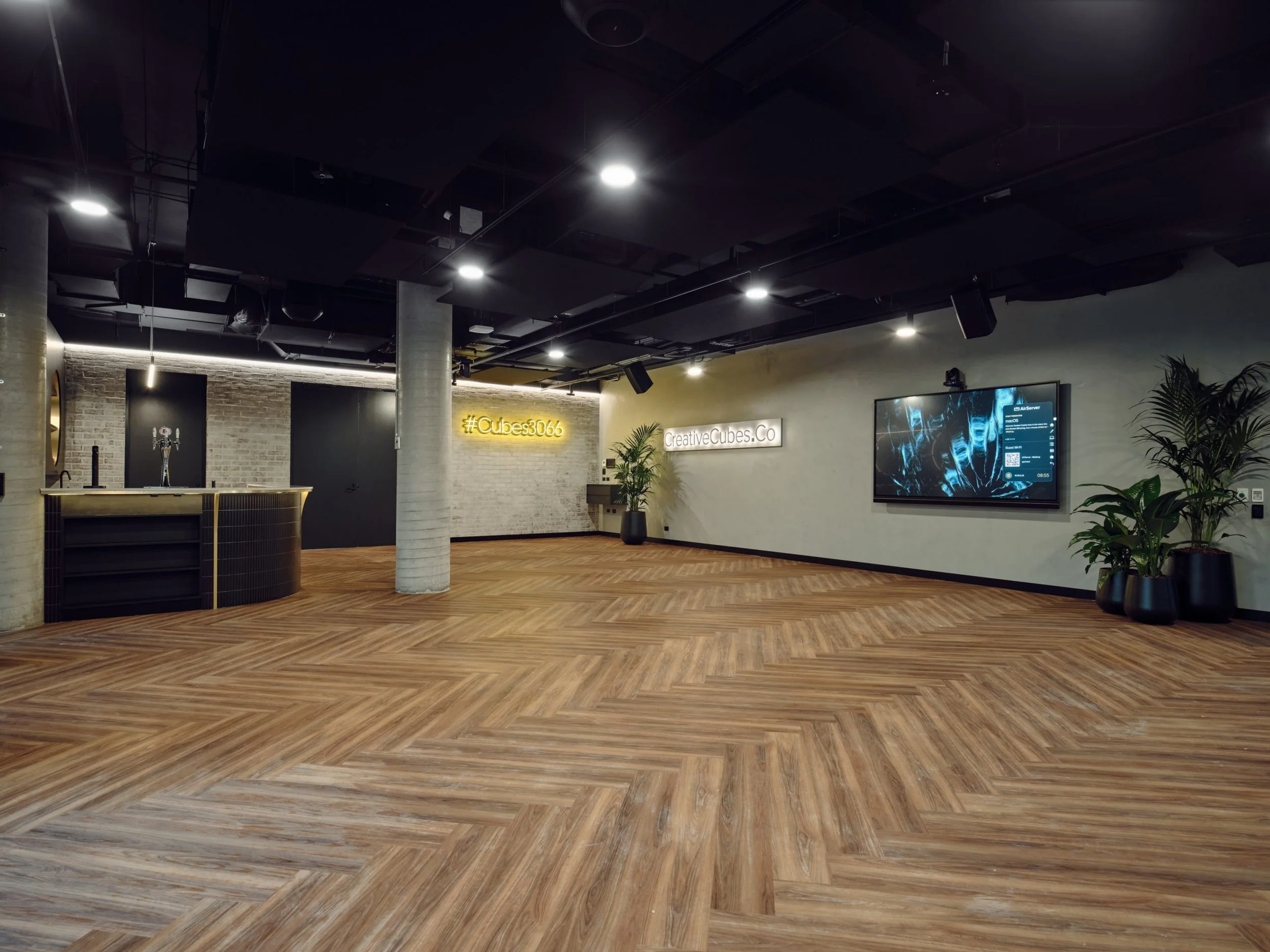 Modern office or conference room with a wooden floor, a large flat-screen monitor on the wall, potted plants, and a light-colored brick and paneled wall with signage, illuminated by ceiling lights.