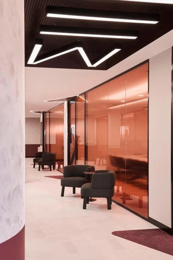 Modern lobby with black armchairs and a reflective reddish-orange wall, illuminated by geometric ceiling lights.