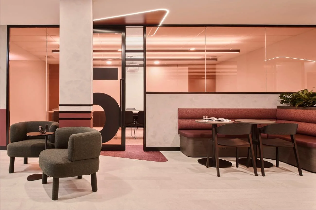 Modern lounge area with two green upholstered chairs, a small table, a curved pink booth with a table and chairs, and a glass wall with black frames, in warm pink lighting.