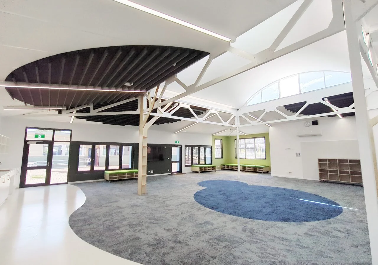 Empty modern classroom or activity room with large windows, green accents, black ceiling panels, and a blue circular rug on a gray carpeted floor.