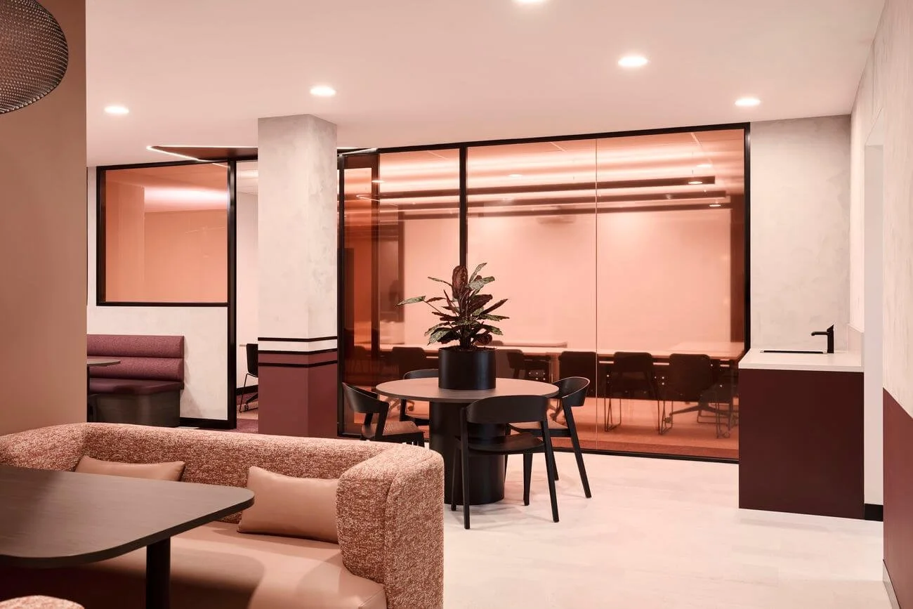 Modern interior with beige sofa, round dining table with black chairs, and a potted plant, separated by glass walls with a pink tint.