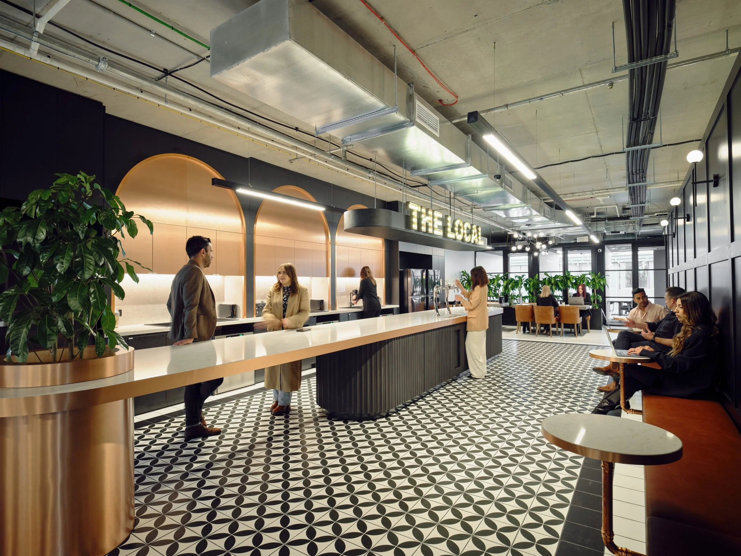 Interior of a modern cafe with a black and white patterned floor, a long white counter, green plants, and several people sitting and standing, with a sign that reads 'The Local' hanging above the counter.