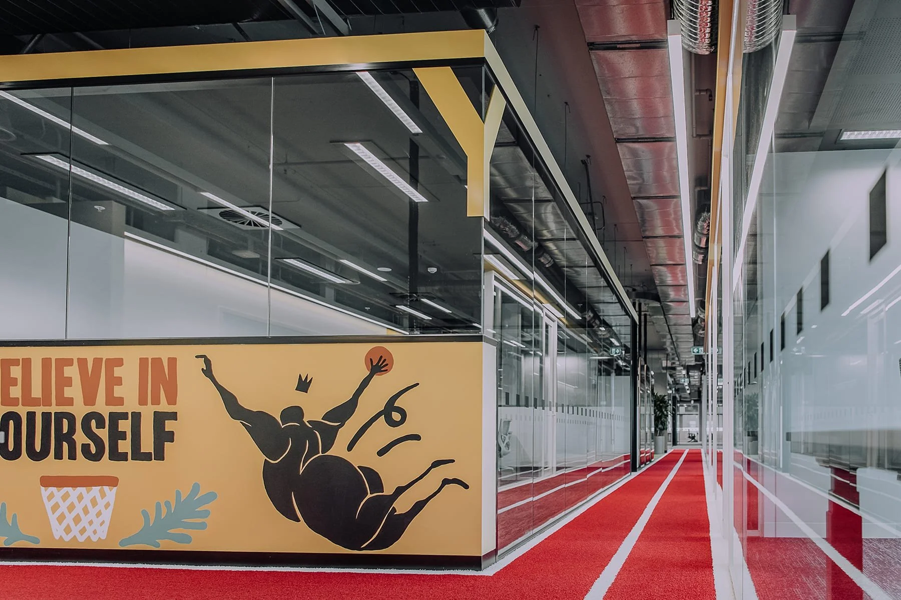 Office hallway with glass walls, red carpet, and motivational poster displaying the text 'Believe in Yourself' and an illustration of a person jumping with arms raised inside a spider silhouette.