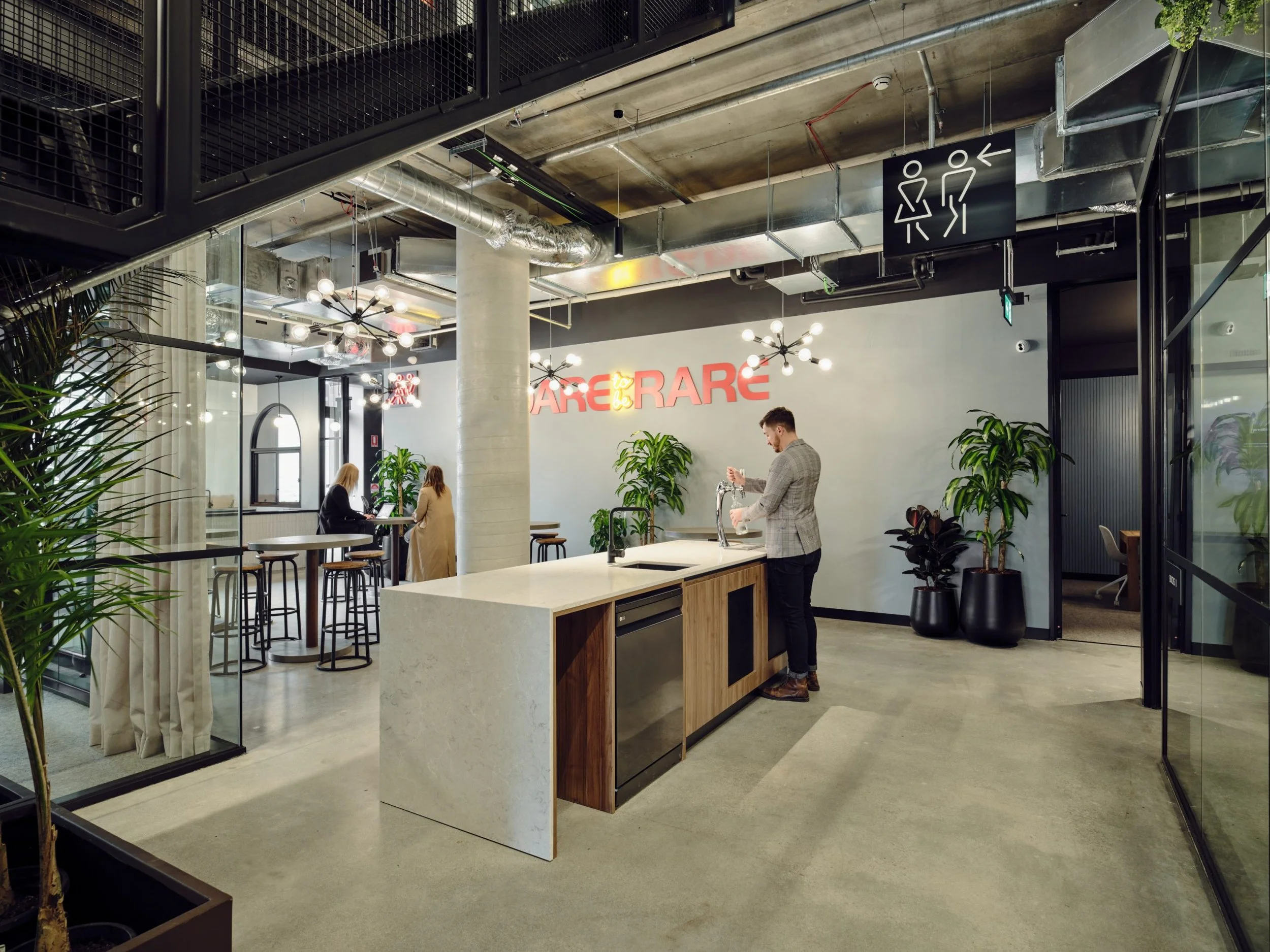 Modern office or coworking space with a kitchen area, plants, and people working. Signage with figures indicates restroom directions.