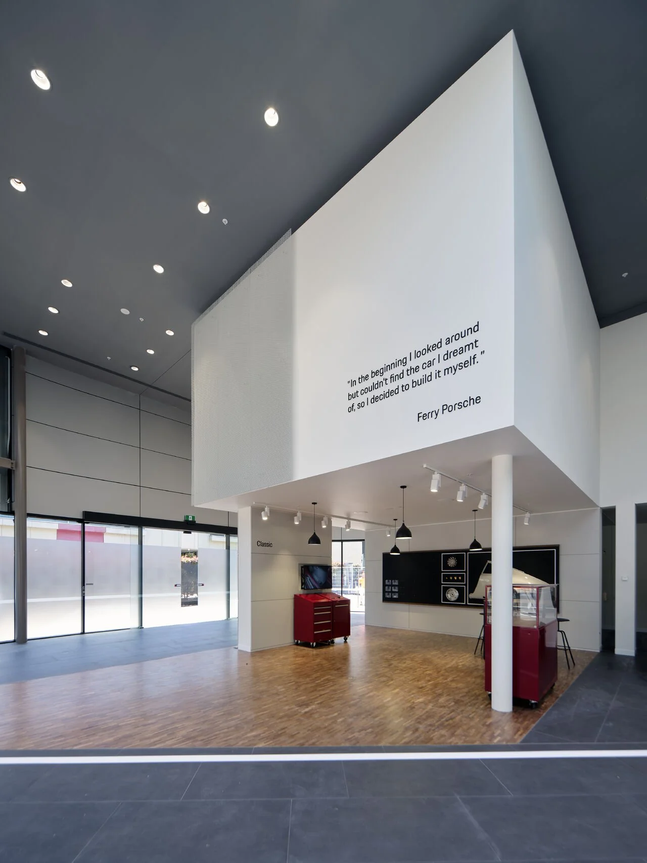 Interior of a modern showroom with a large white wall featuring a quote by Ferry Porsche, displaying a sleek, minimalistic design with black pendant lights, red display counters, and a wooden floor.