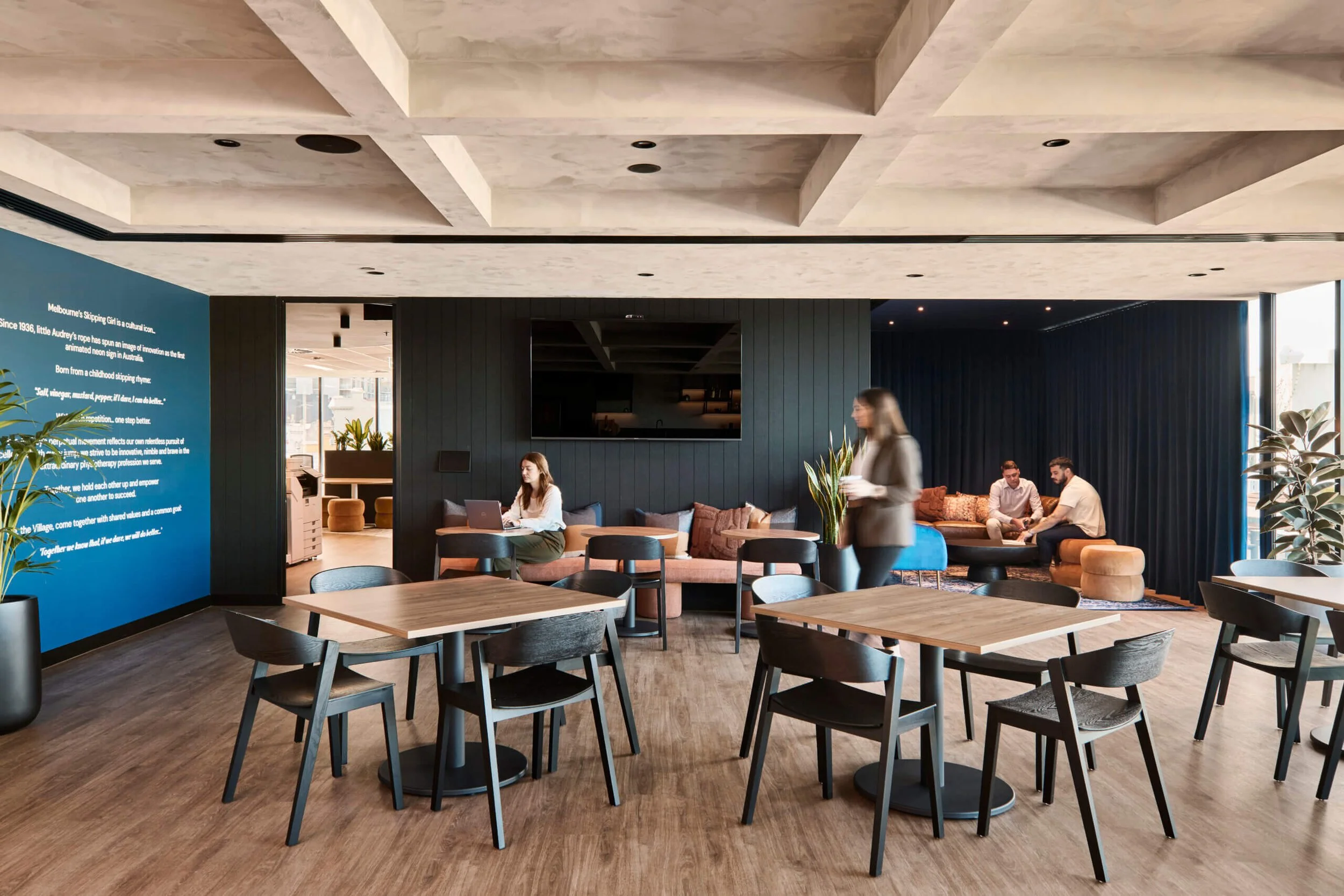 Modern, stylish lounge area with wooden tables and black chairs, a blue wall with white text, large windows, and several people sitting and walking around.