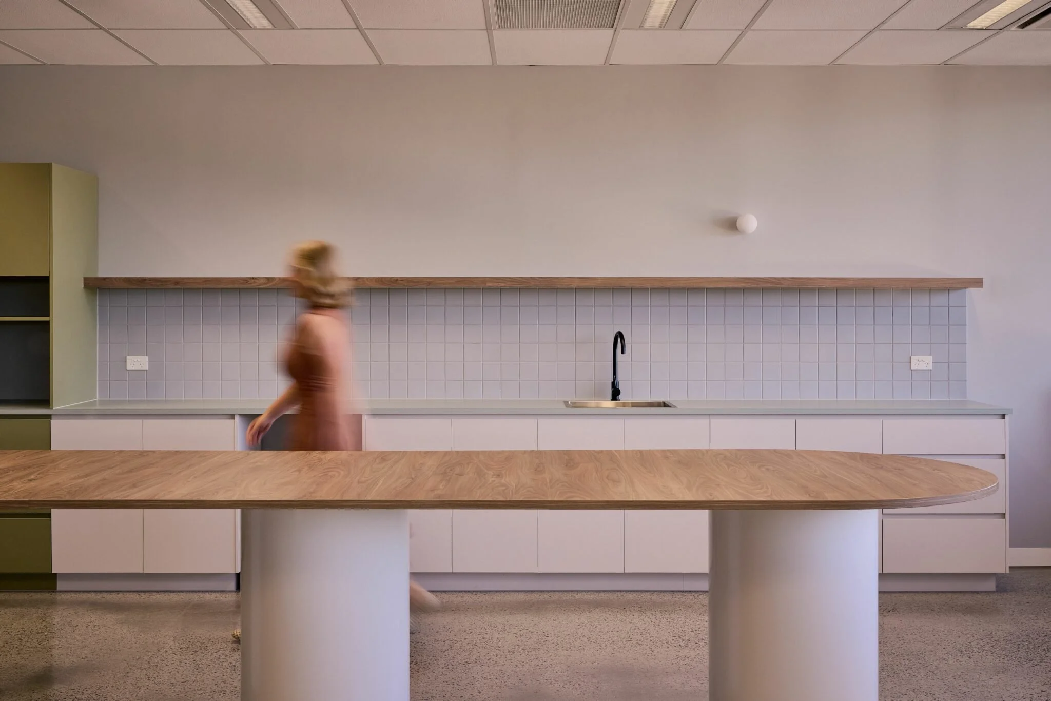 A person walking in a modern kitchen with a white tiled backsplash, light-colored cabinetry, and a wooden countertop. The person is blurred, indicating motion.