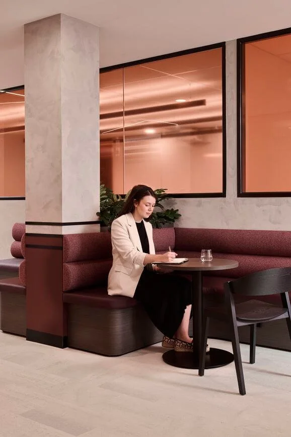 A woman sitting at a round table in a modern cafe or restaurant, writing in a notebook with a glass of water beside her. The setting has mauve and beige tones with some plants and large windows in the background.