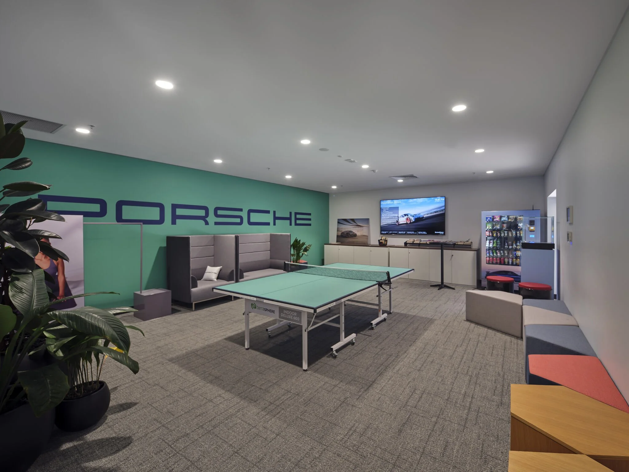 A seating area in a showroom or lounge with a cardboard ping pong table, a large Porsche logo on a green wall, a vending machine, and modern chairs and plants.