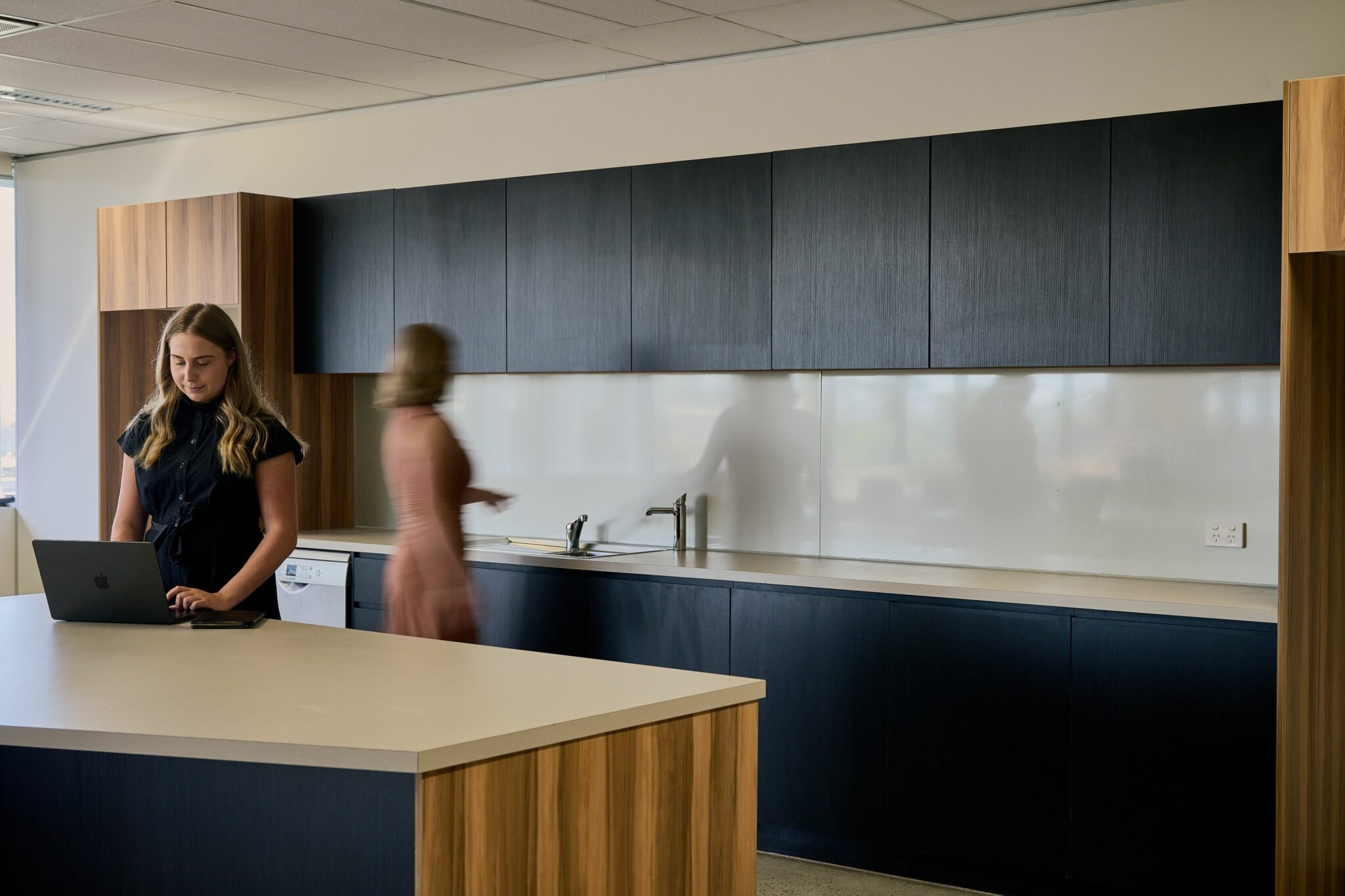 Interior view of a modern office kitchen with dark blue cabinets, a white backsplash, a sink, and a light-colored countertop. A woman with long blonde hair, wearing a black shirt, is working on a laptop at the counter. Blurred figures are walking in 