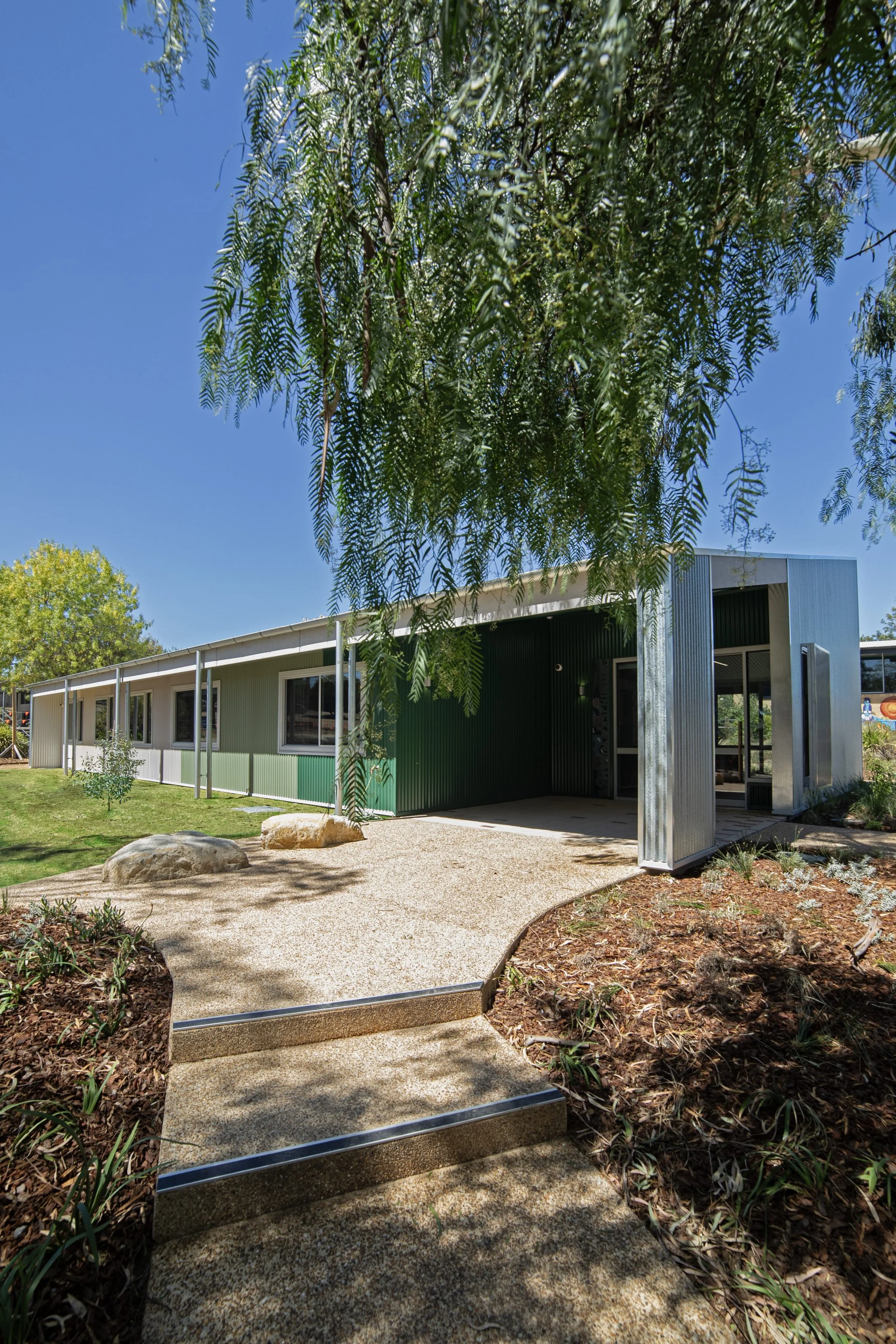 Modern building with corrugated metal siding, large windows, and a curved walkway leading to the entrance. Tall trees and a landscaped garden with rocks and mulch surround the area.