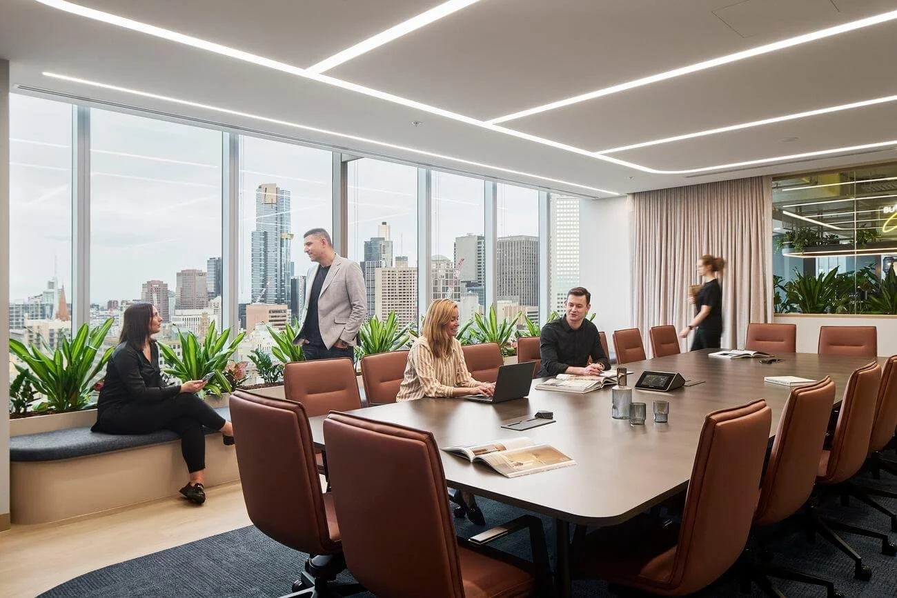 Modern conference room with a large table, brown chairs, and a city skyline view through floor-to-ceiling windows. Five people are inside: one woman sitting on a window ledge using her phone, another woman working on a laptop, a man with papers, and 