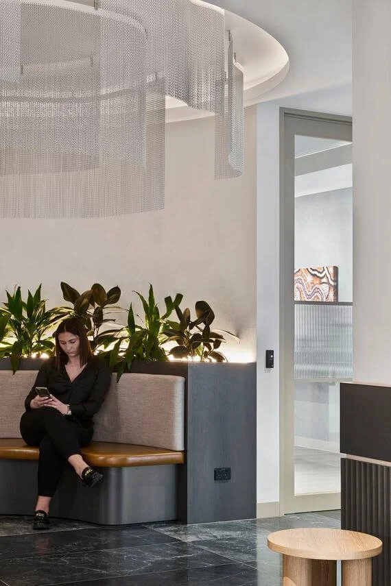 A woman sitting on a built-in beige and brown bench indoors, looking at her phone, with green plants behind her and a modern interior design.
