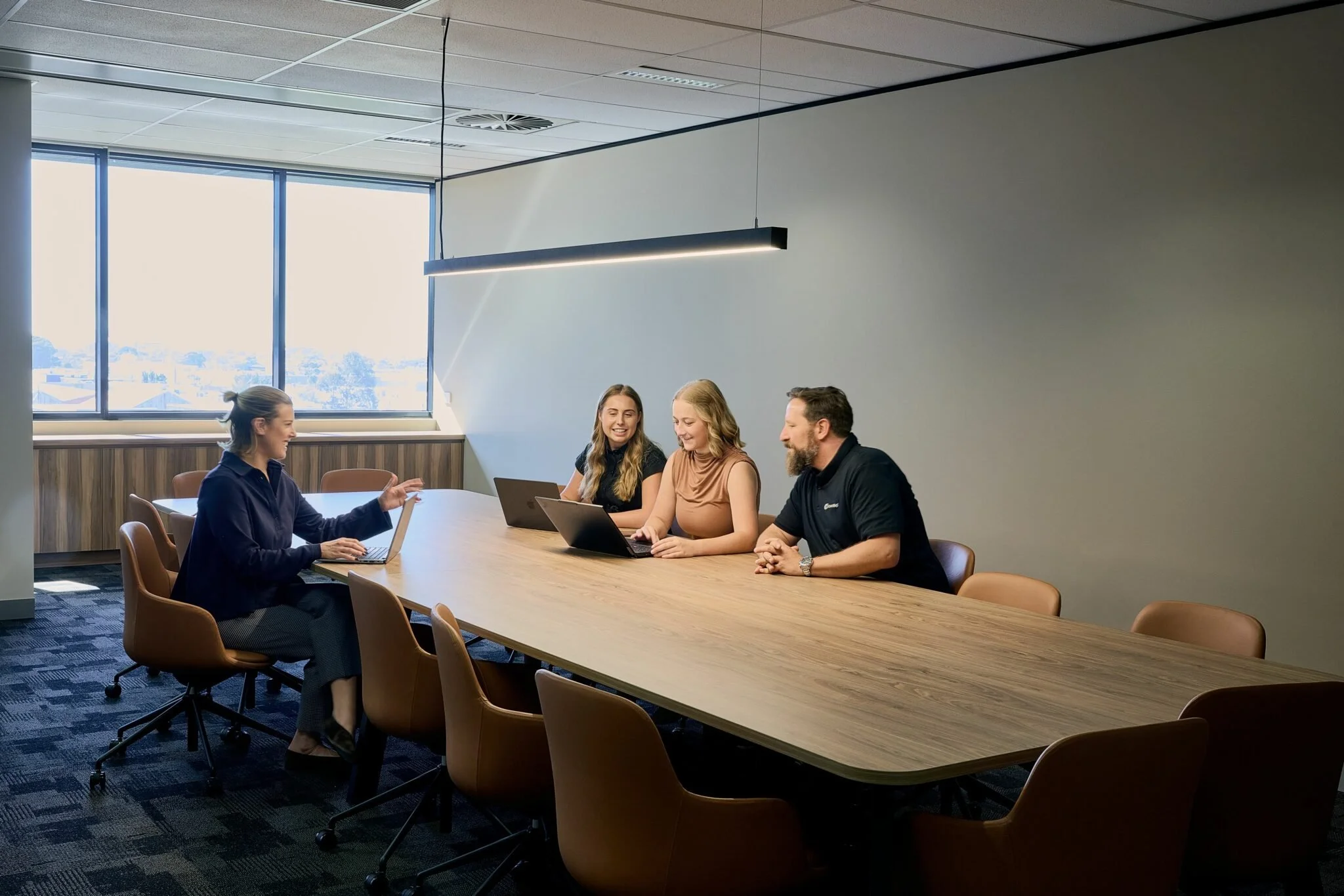 Four people sitting around a conference table in a modern office, engaged in discussion, with laptops open in front of them.