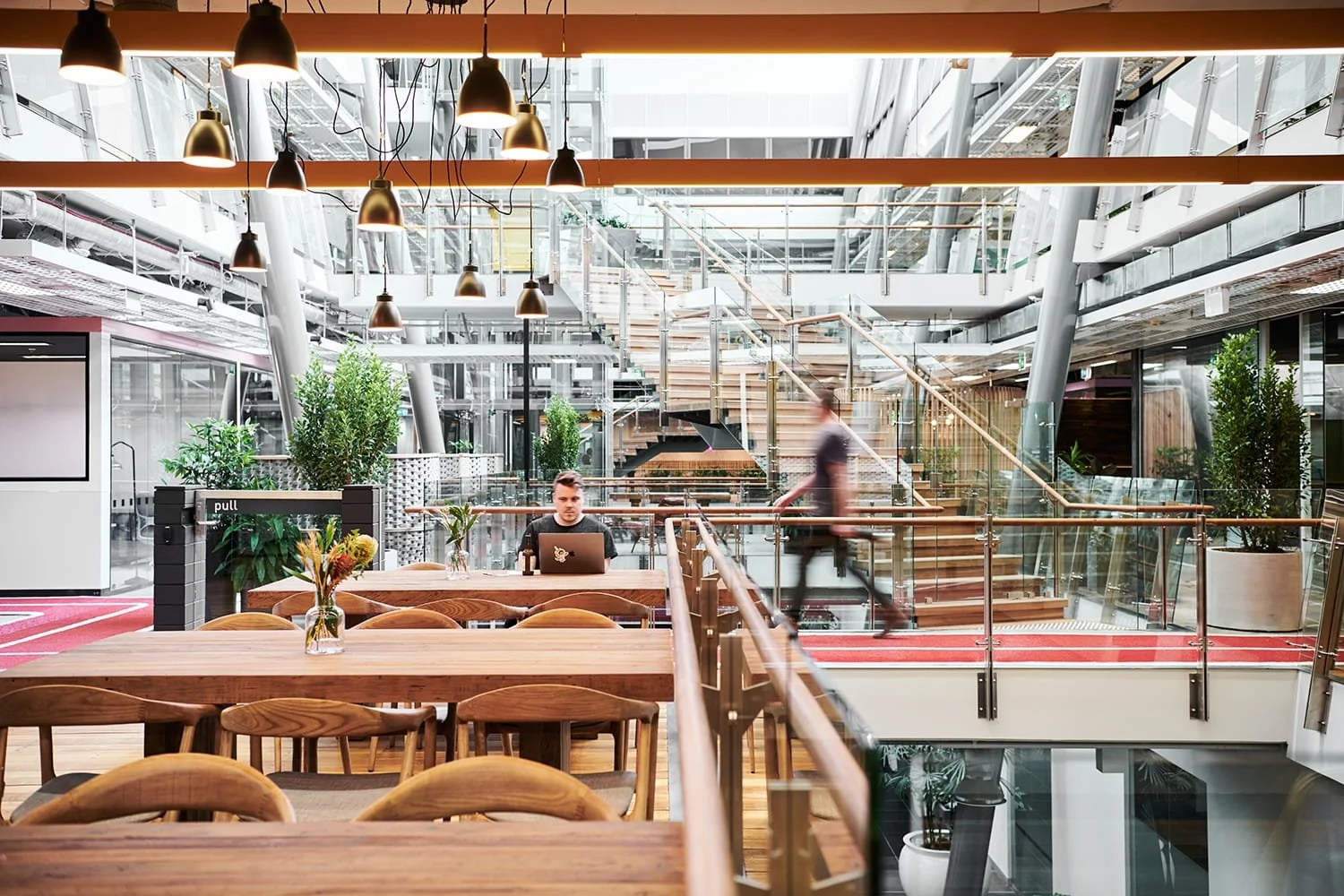 Interior of a modern, multi-level office or co-working space with wooden furniture, plants, stairs, and glass walls, featuring a person working on a laptop at a table and another person walking.