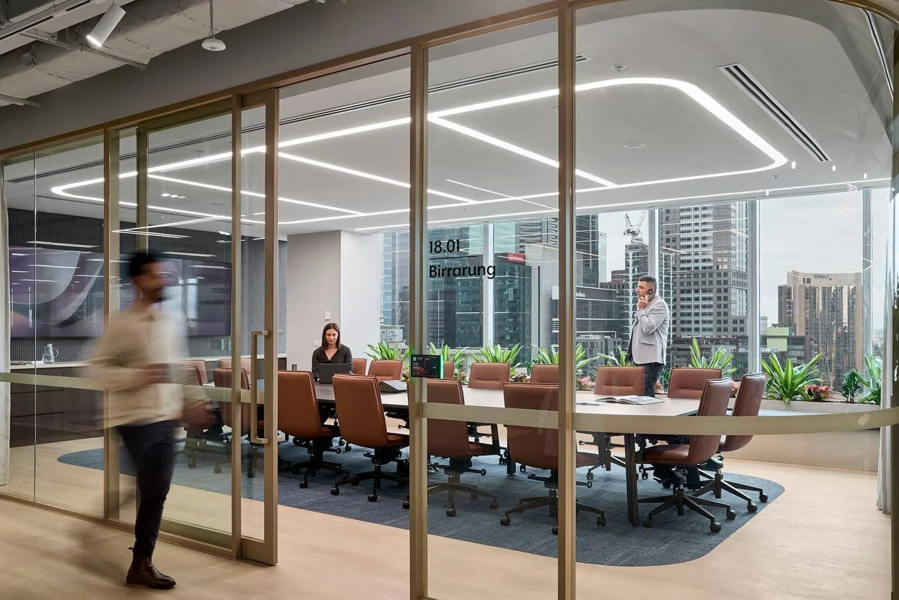 Modern office conference room with glass walls, a large table, and brown chairs. Two people are inside; one is working on a laptop, and the other is on the phone. A blurred person is walking past outside the room. City skyscrapers are visible through