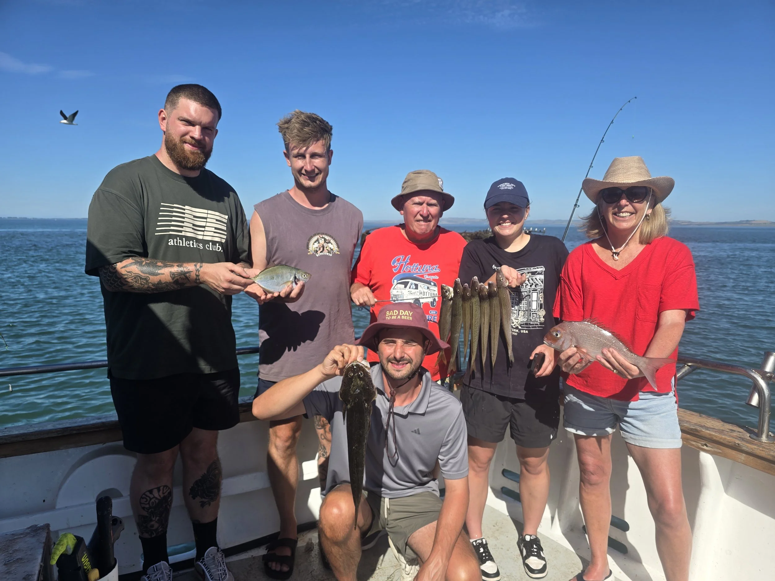 Group of six people on a boat holding fish with a lake or ocean in the background and a bird flying in the sky.