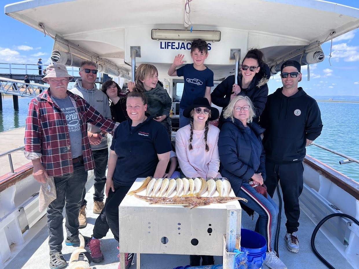Group of people on a boat with a table of fish in front of them, posing for a photo with water and a pier in the background.