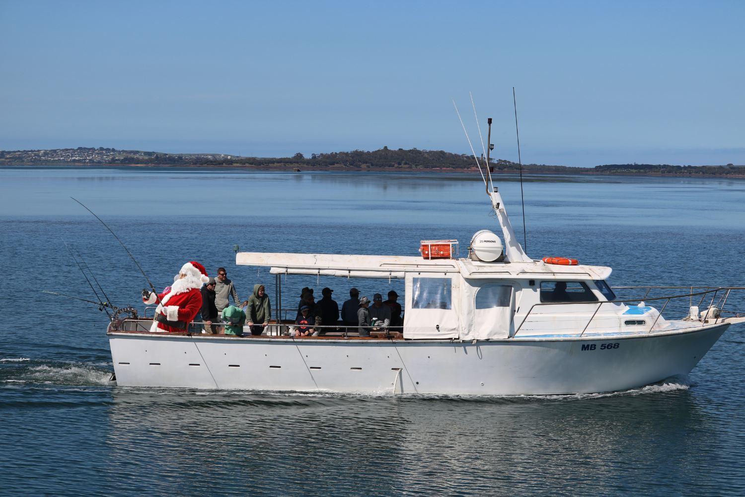A boat on the water with a person dressed as Santa Claus handing out gifts to people on the boat, with a scenic shoreline in the background.