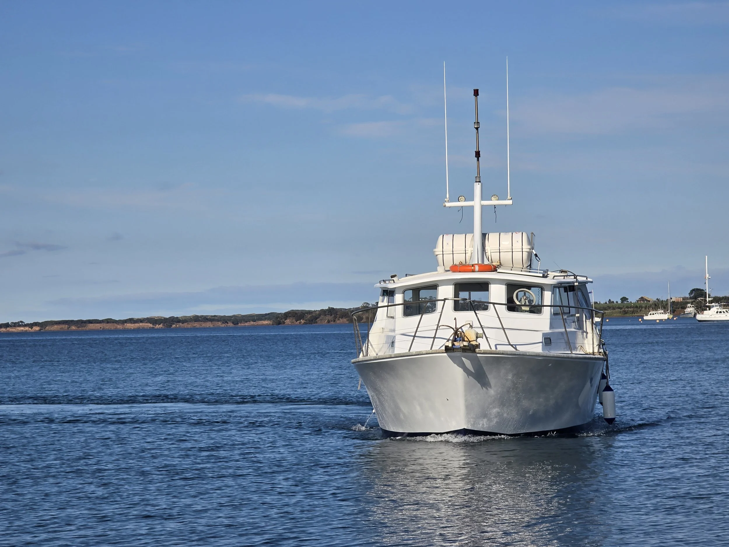A white boat with radar and antenna equipment on top is moving through a body of water, with other boats visible in the background under a clear blue sky.