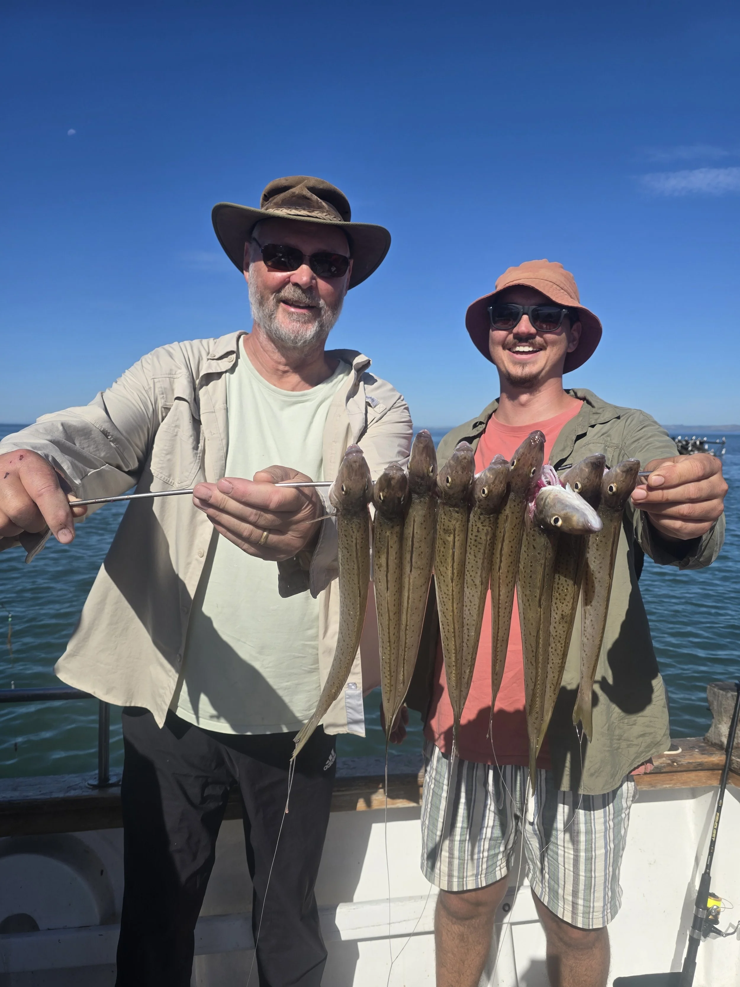 Two men smiling and holding a string of freshly caught fish on a boat, with clear blue sky and ocean in the background.