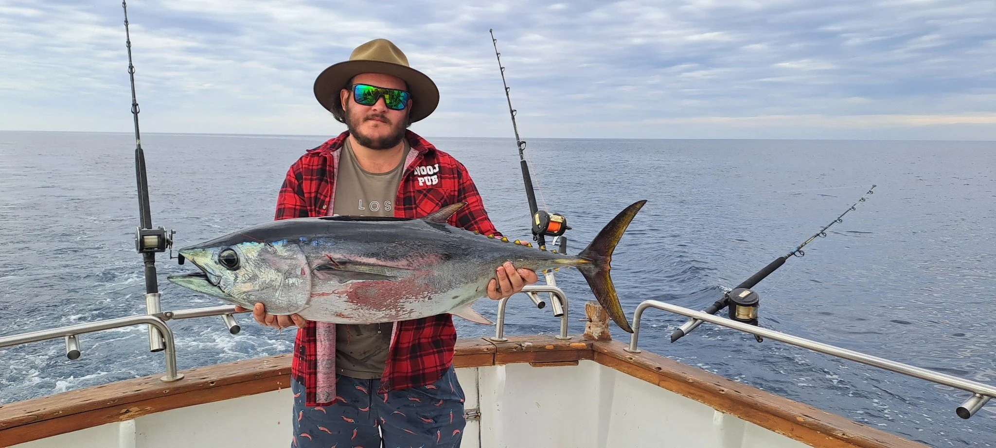 A man holding a large fish on a boat at sea, with fishing rods and water in the background.