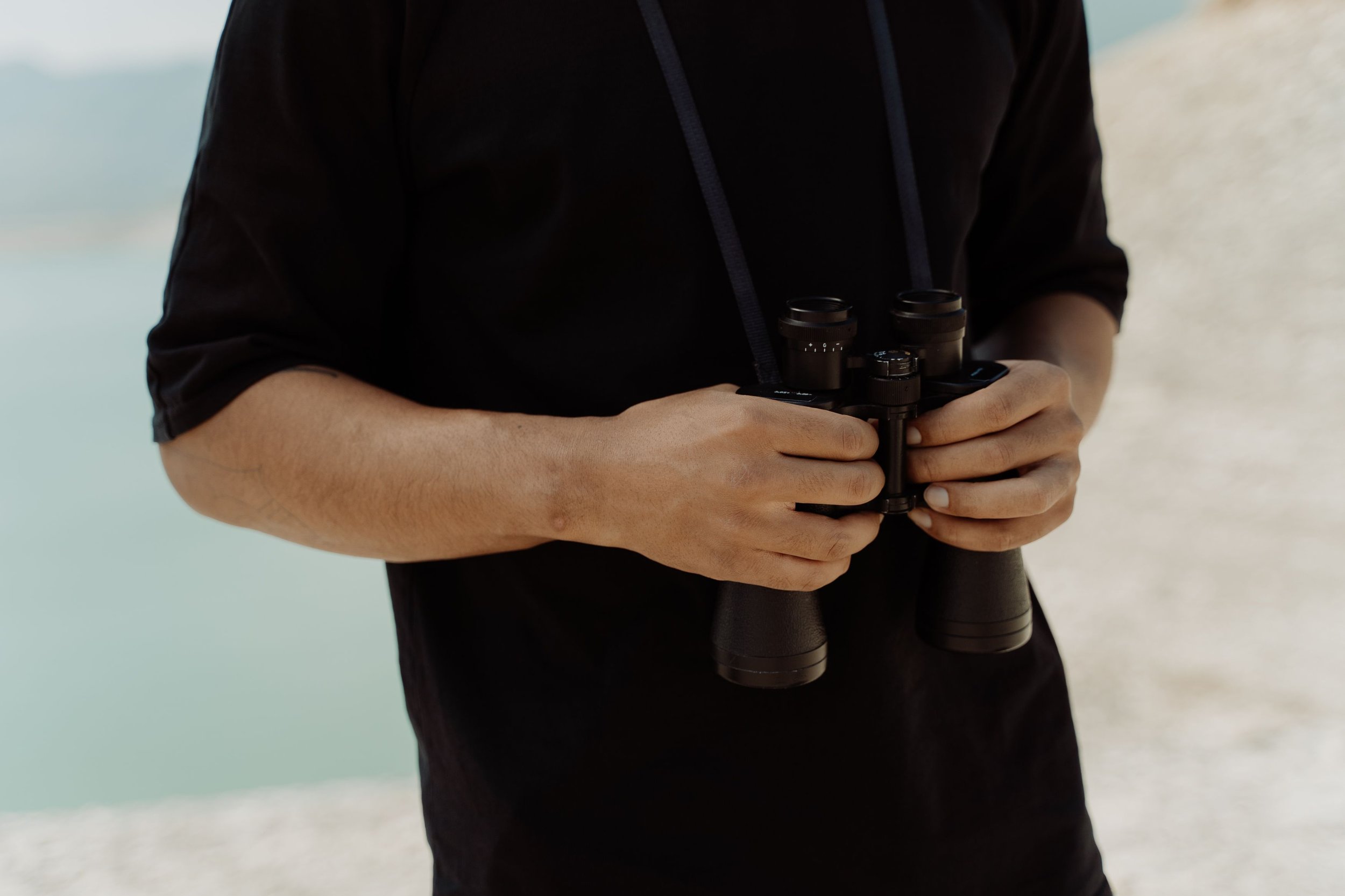 Person holding binoculars on a beach with a sandy shoreline and water in the background.