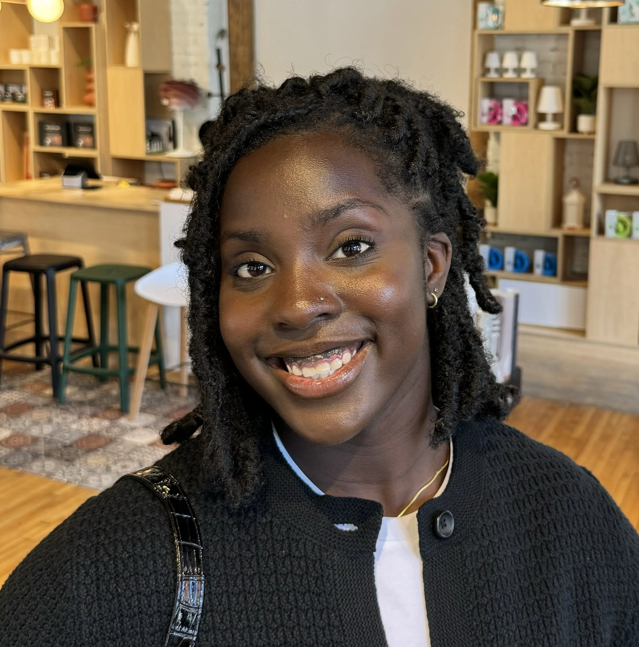 Close-up of a smiling woman with curly black hair, wearing a black textured sweater, gold earrings, and a gold necklace, in a cozy indoor setting with wooden shelves and stools in the background.