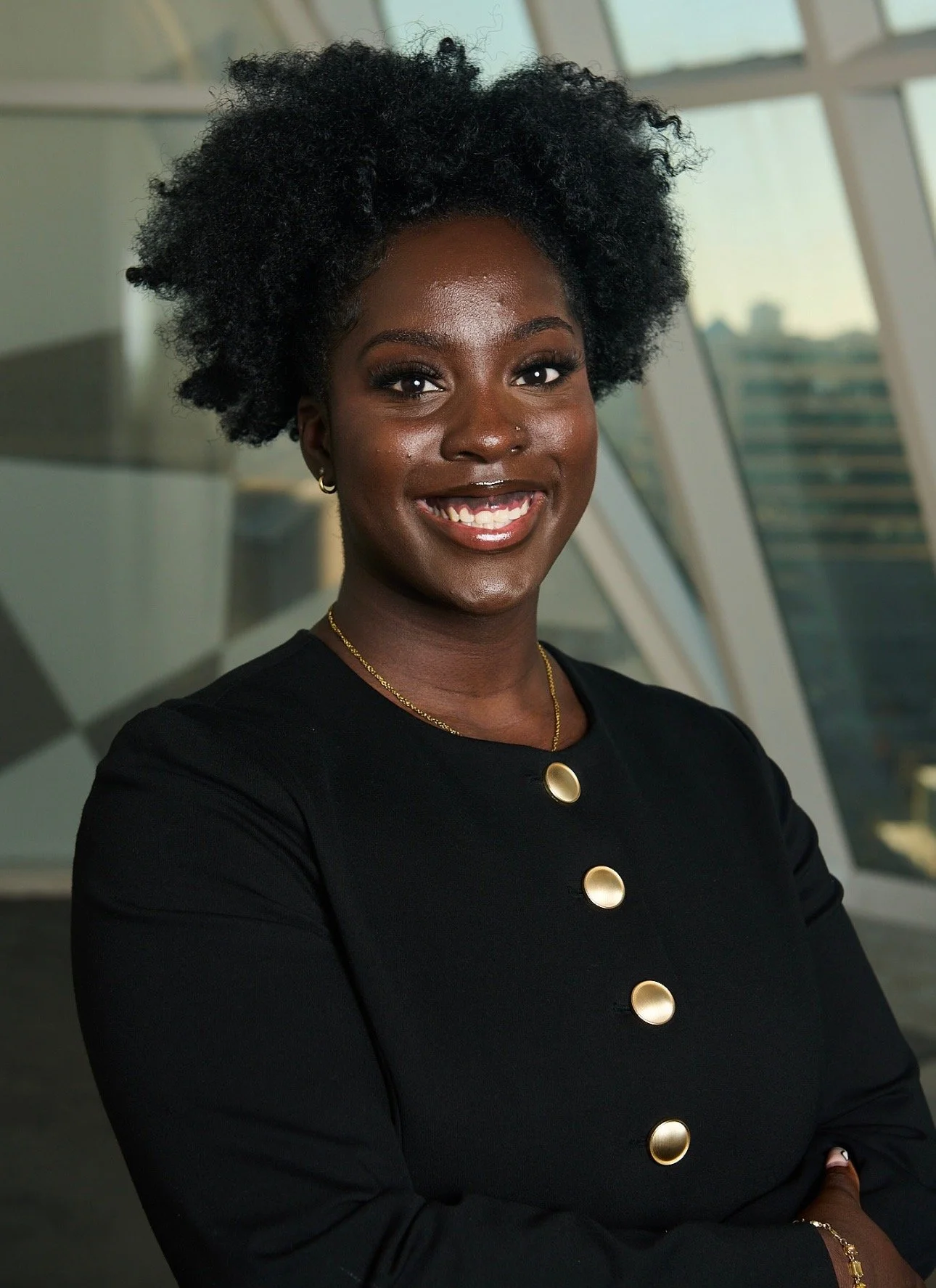 Portrait of a smiling Black woman with curly hair, wearing a black dress with gold buttons and jewelry, standing indoors near large windows.