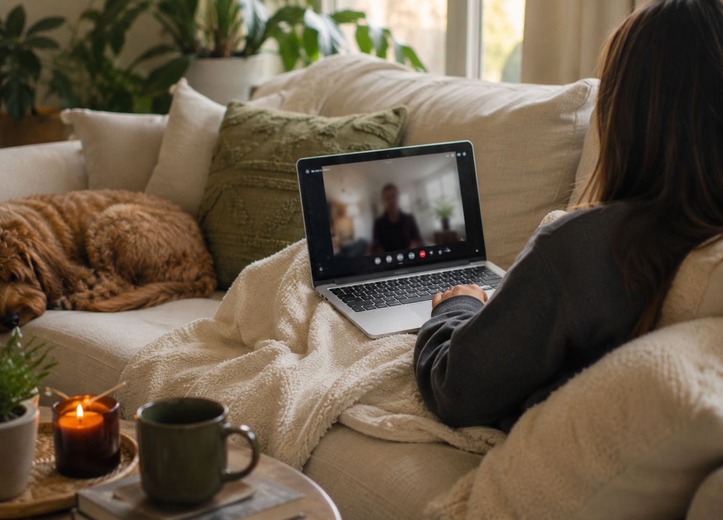 Person lying on a sofa using a laptop, talking to someone on video call, with a dog sleeping next to them; cozy living room setting with plants, books, candles, and a window in the background.