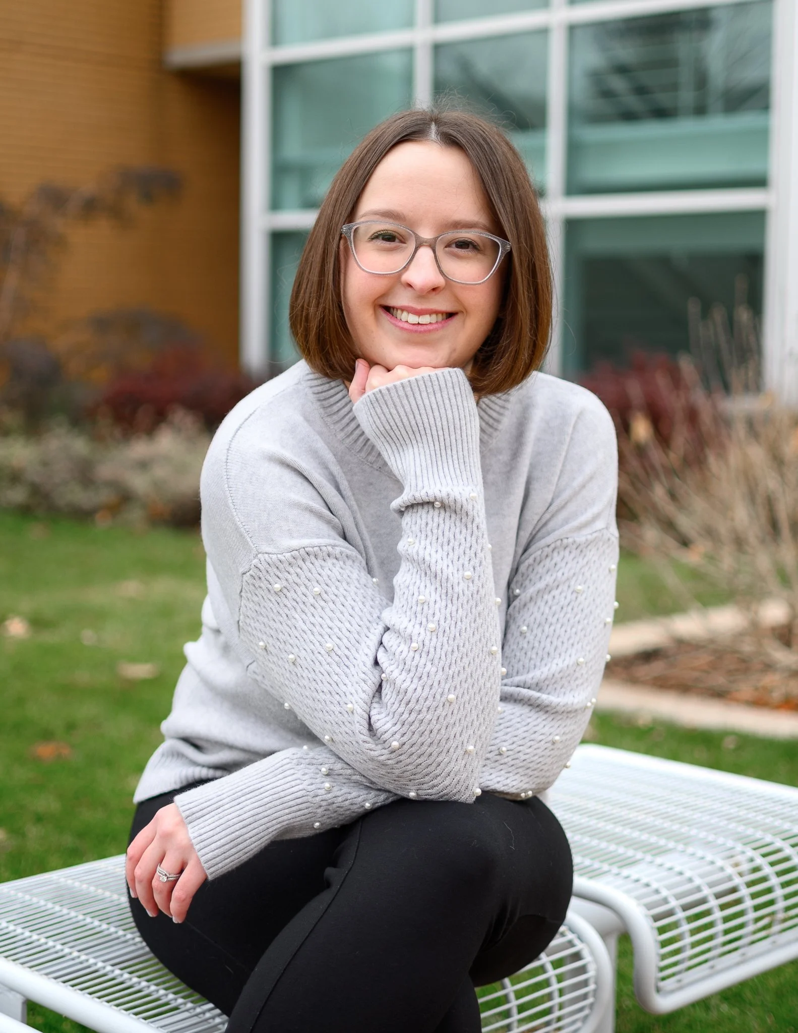 A woman with shoulder-length brown hair, wearing glasses, a gray sweater with pearl embellishments, and black pants, sitting on a white metal bench outdoors near a modern building with large glass windows and a brick wall.