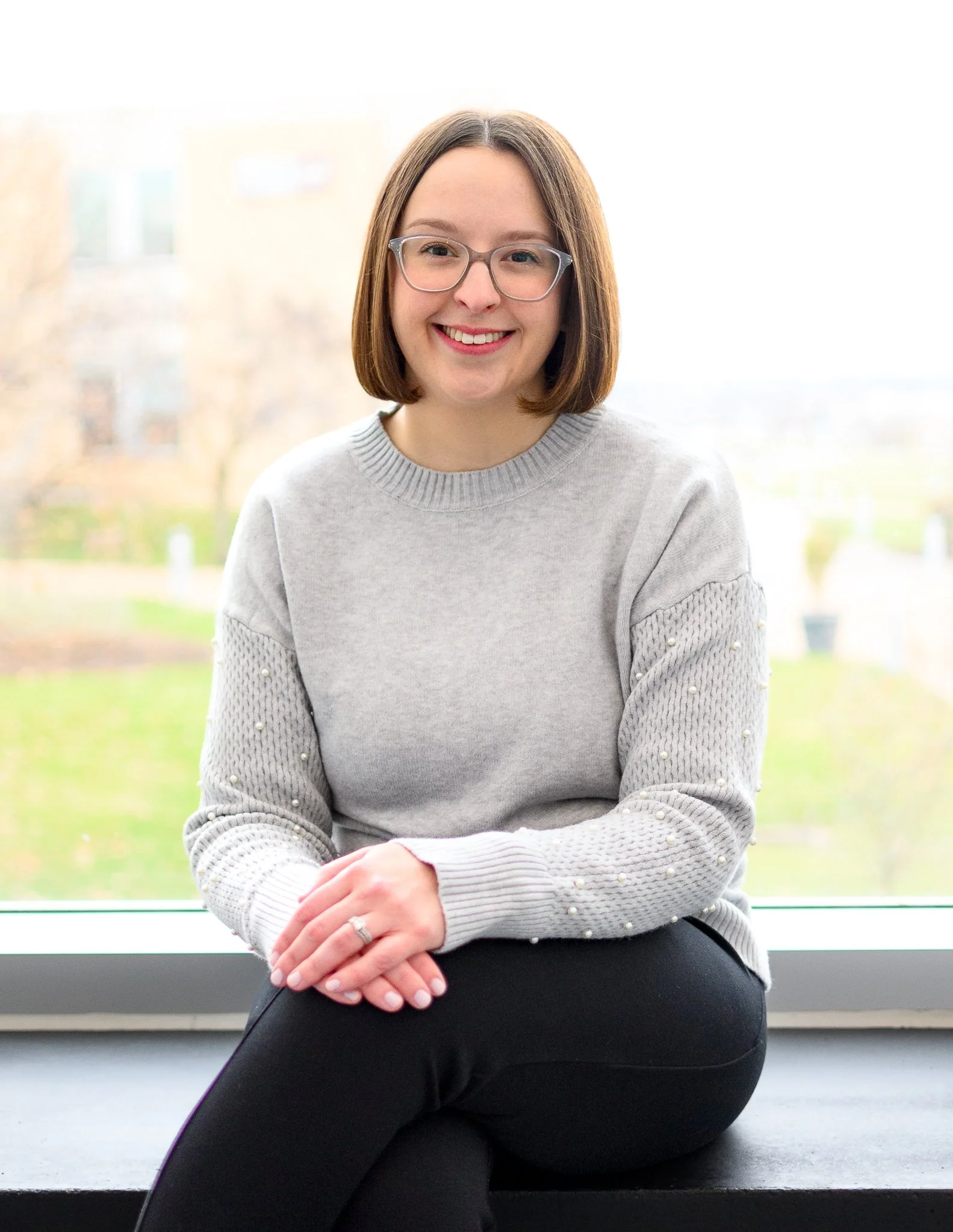 A woman with shoulder-length brown hair, glasses, and a warm smile sitting indoors near a bright window with a blurry outdoor scene behind her. She is wearing a light gray sweater with pearl embellishments on the sleeves and black pants, and she is sitting with her hands crossed in her lap.