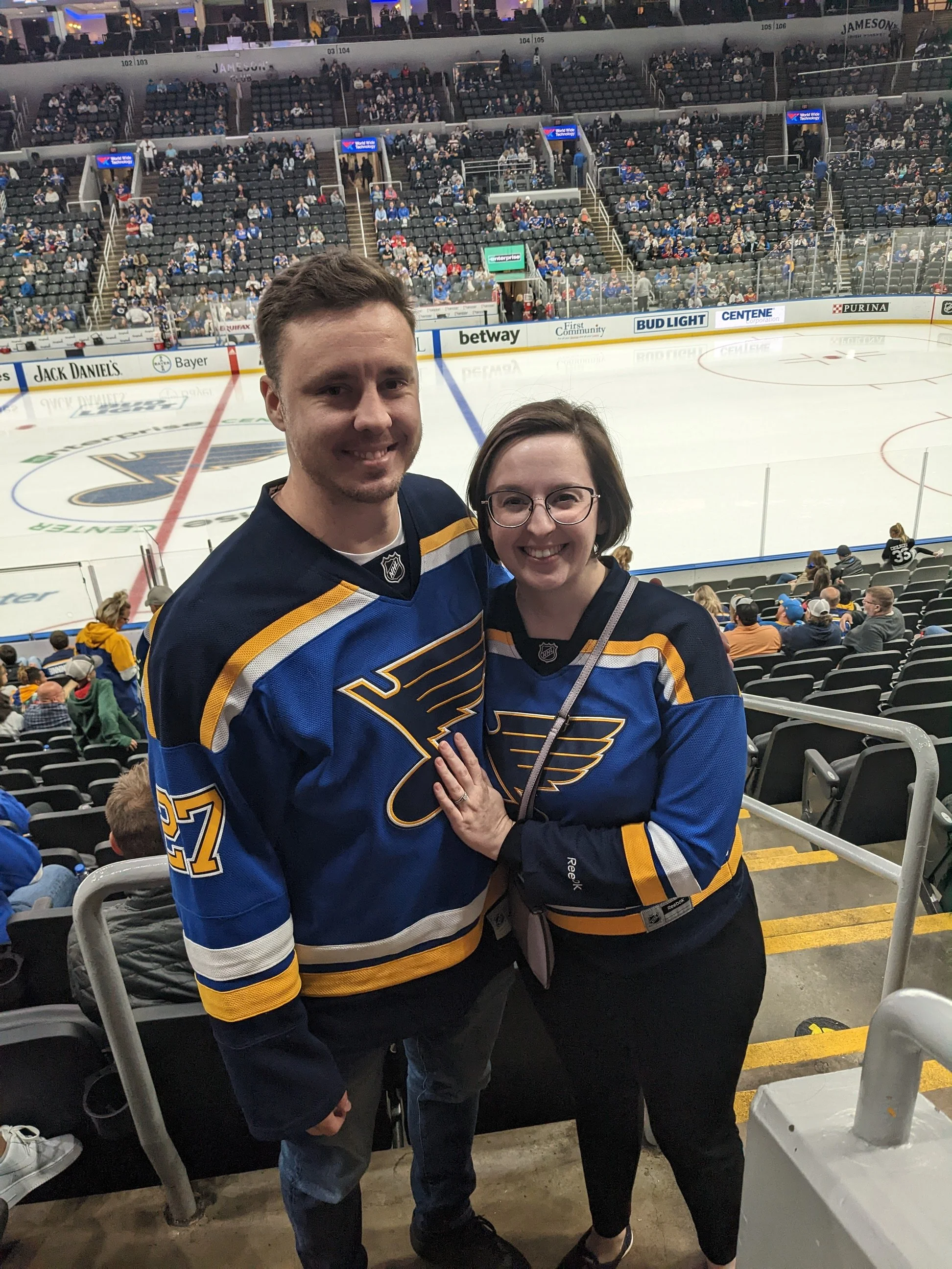 Two people wearing St. Louis Blues hockey jerseys standing together in an ice hockey arena with a crowd in the background.