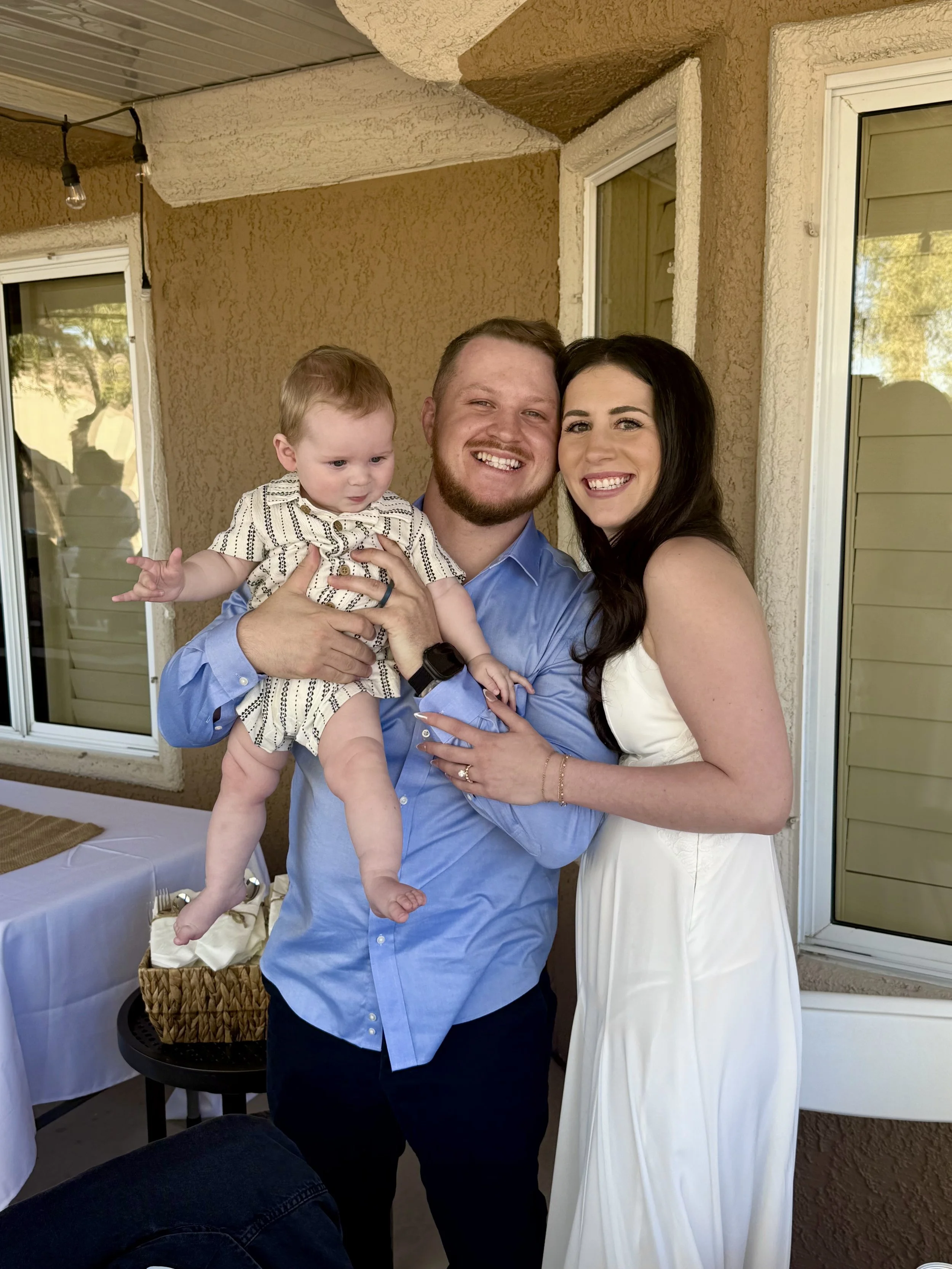 A happy couple smiling with a baby on a patio, with a beige stucco exterior wall, windows, and a small table with a white tablecloth in the background.