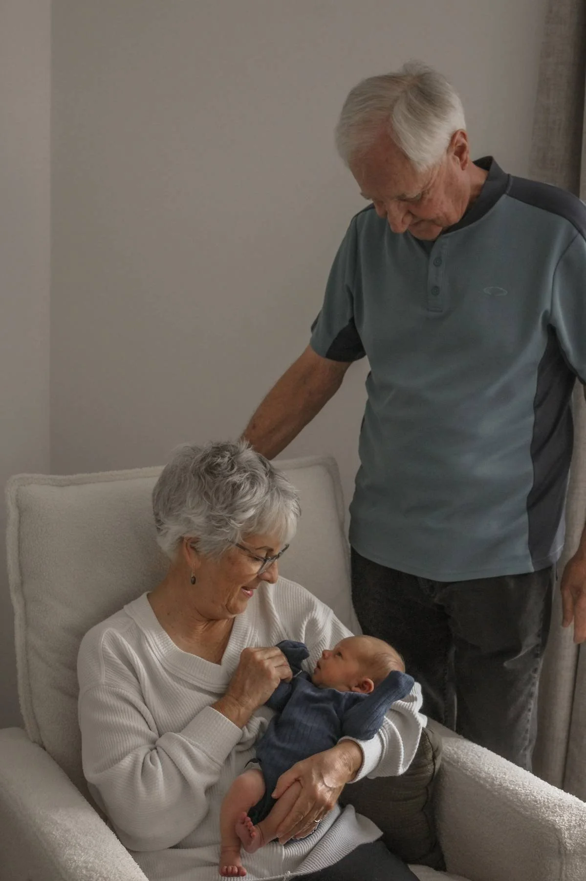 An elderly woman with gray hair and glasses sitting on a white upholstered armchair holding a newborn baby or infant in her lap. An elderly man with gray hair in a gray polo shirt stands beside her, leaning over slightly and looking down at the baby. The background shows a plain wall with a window curtain.