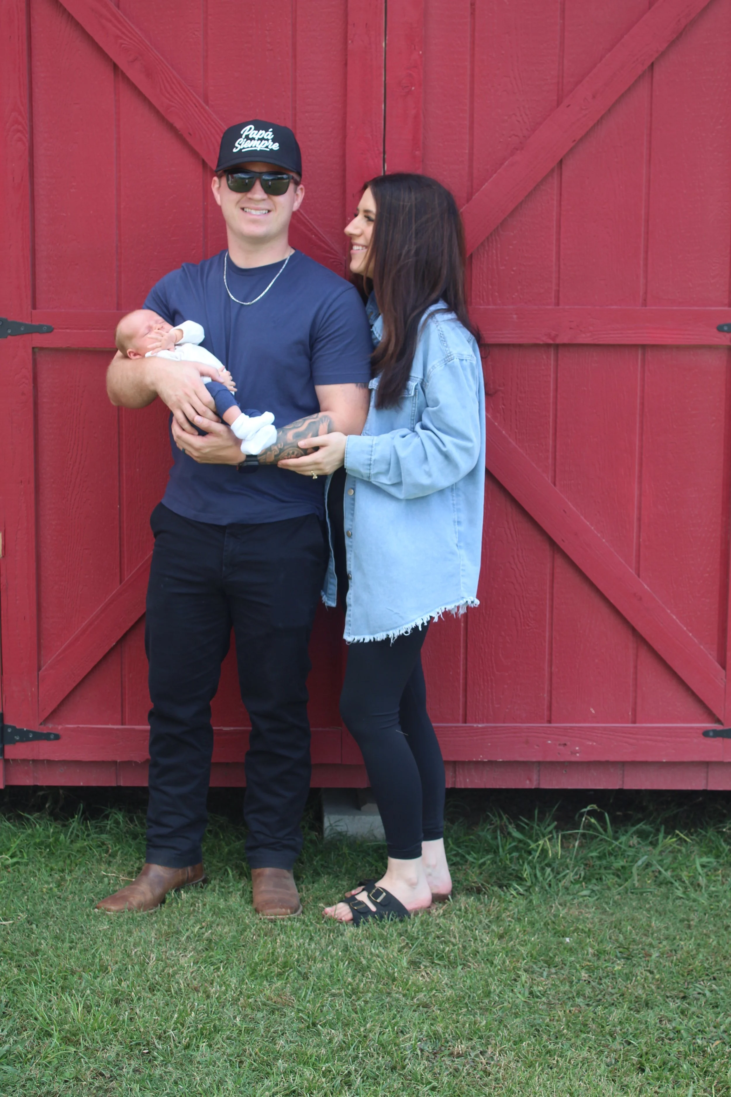A man and woman standing in front of a large red barn door, holding a sleeping baby, all smiling and dressed casually.