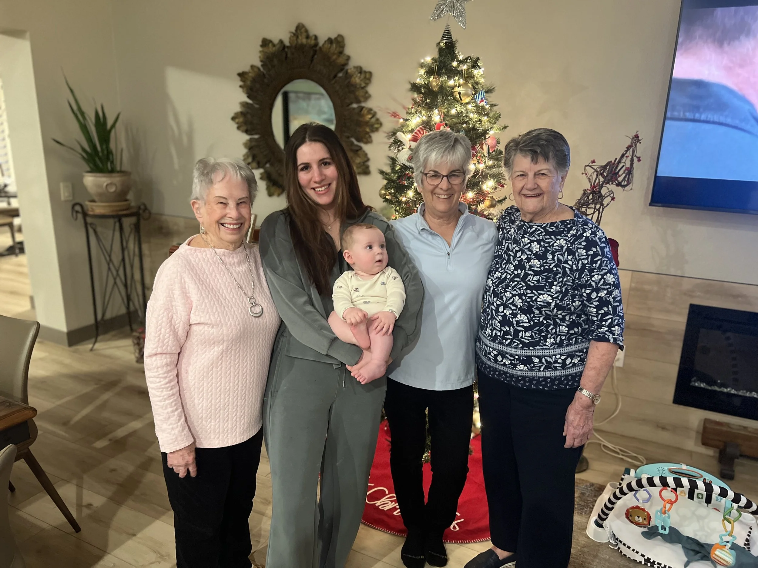 Five women and one baby are standing in front of a Christmas tree, smiling, inside a home decorated for Christmas.