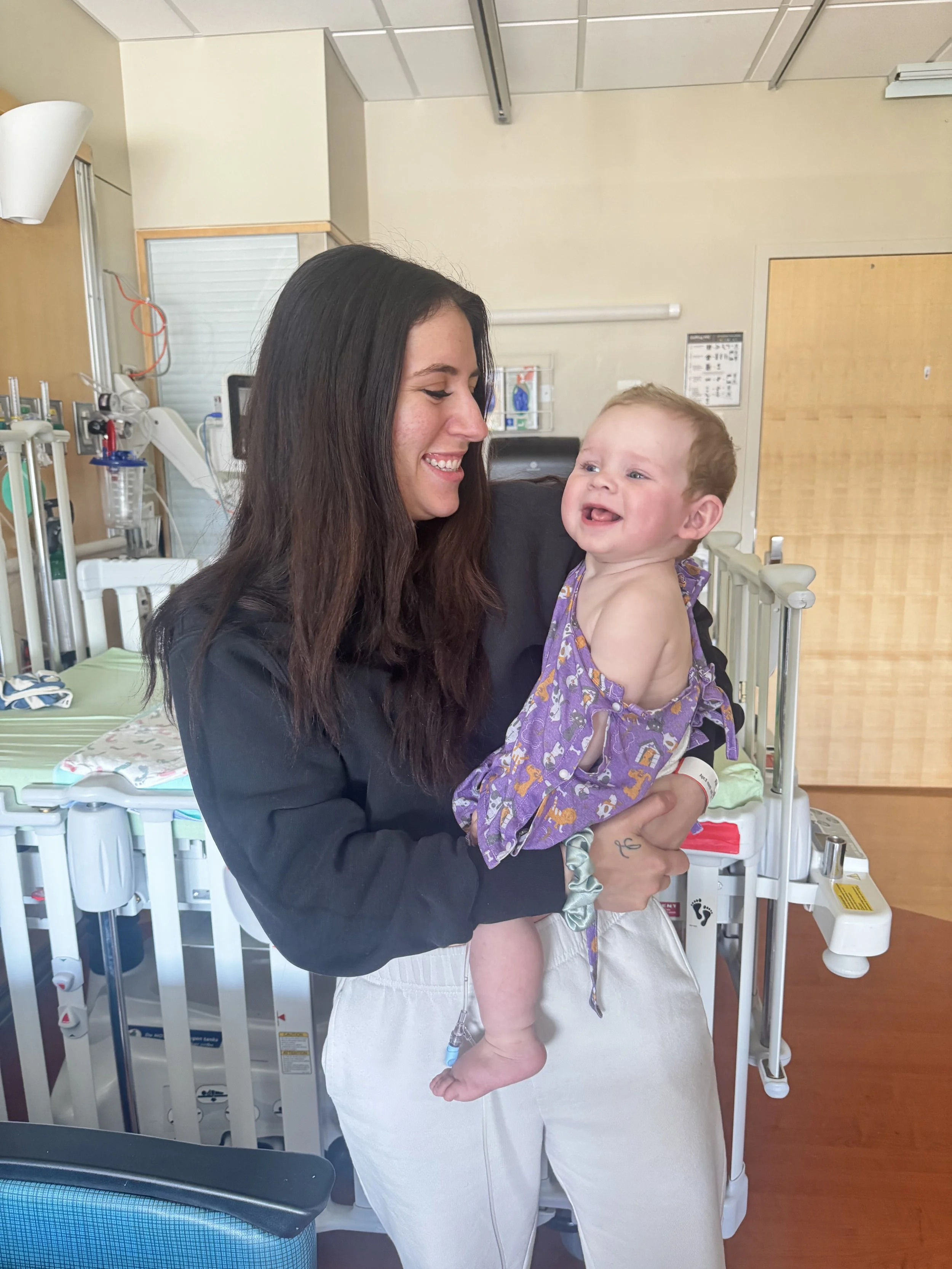 A woman holding a laughing baby in a hospital or clinic room, with medical equipment and a hospital bed in the background.