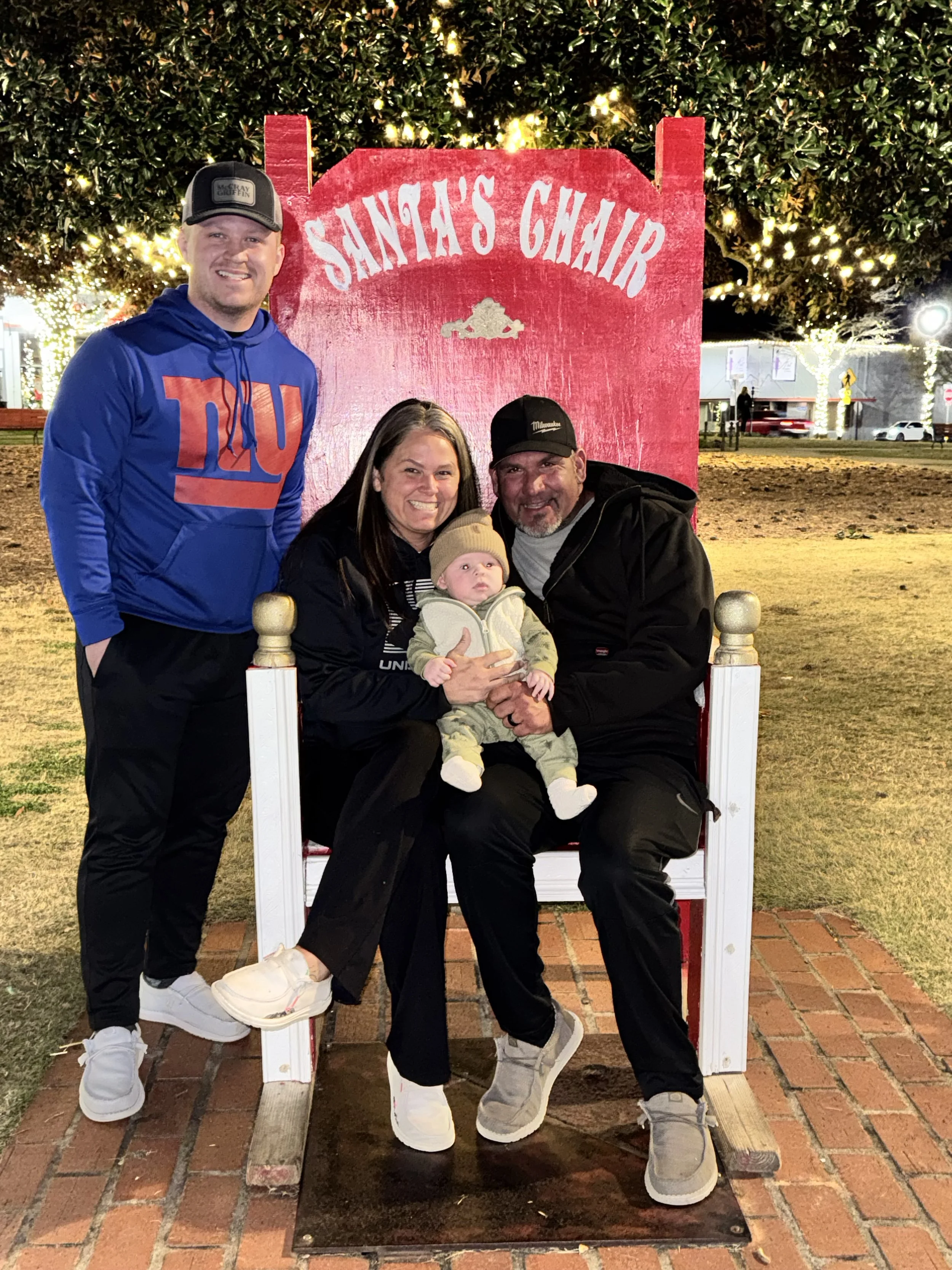 A family of four posing on a Christmas-themed display seat labeled 'Santa's Chair' at night, decorated with lights and Christmas trees in the background.