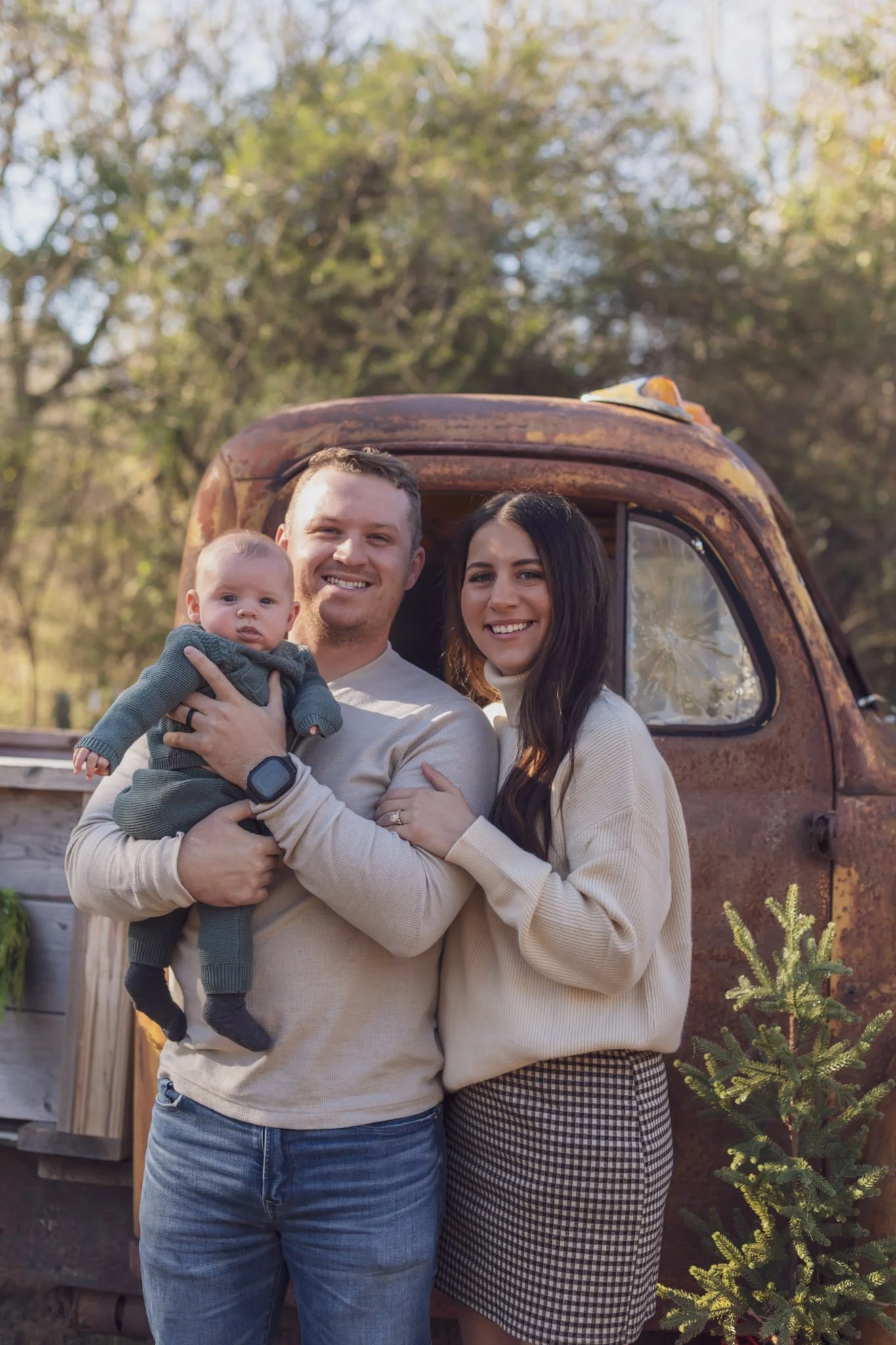A family of three, including a man, woman, and a baby, standing outdoors in front of an old rusty truck and trees with yellow and green leaves.
