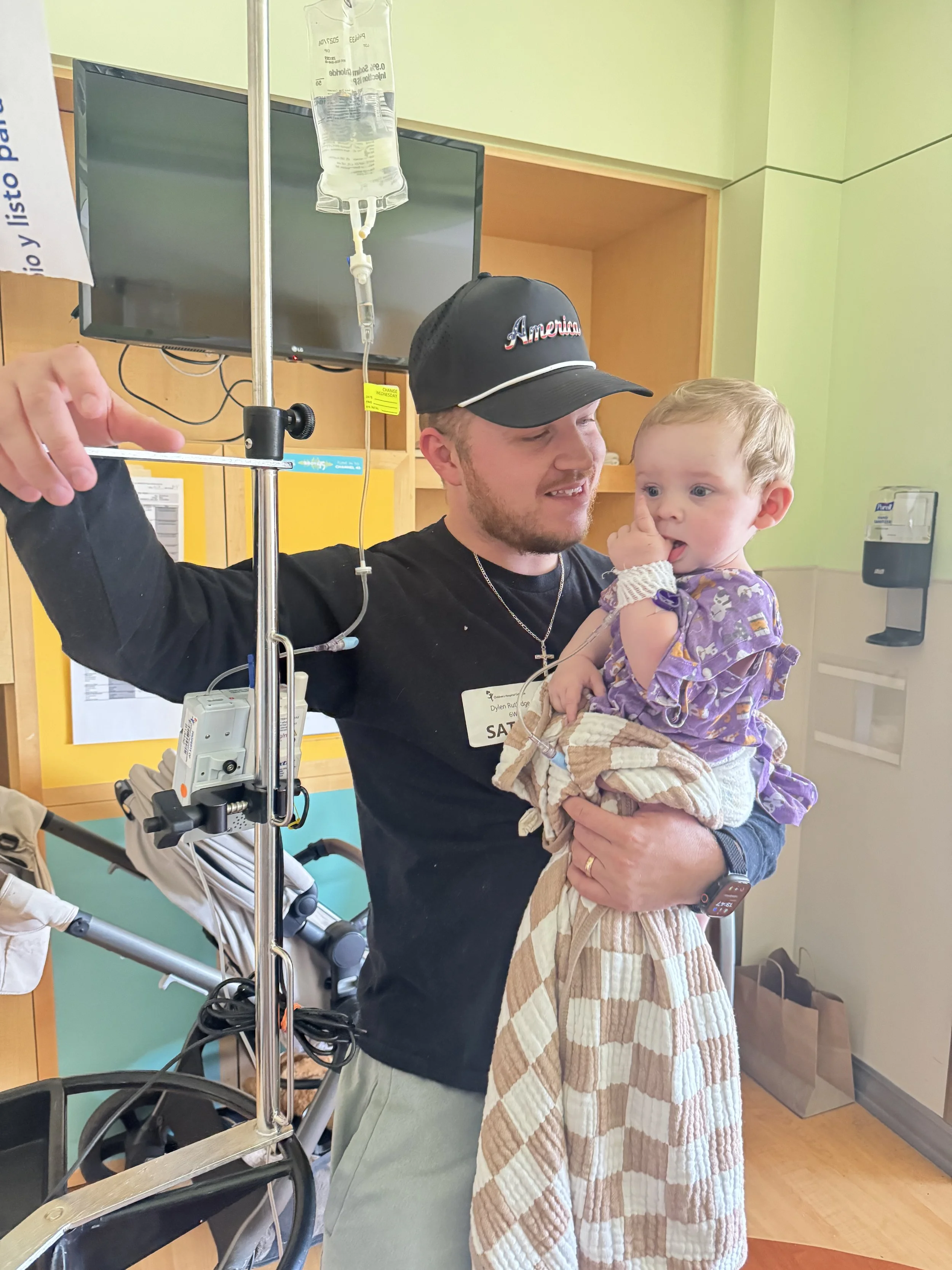 A man in a black shirt and cap holding a young girl in a hospital setting. The girl is wearing a purple dress and a beige quilted blanket. An IV bag hangs above, and medical equipment is visible in the background.