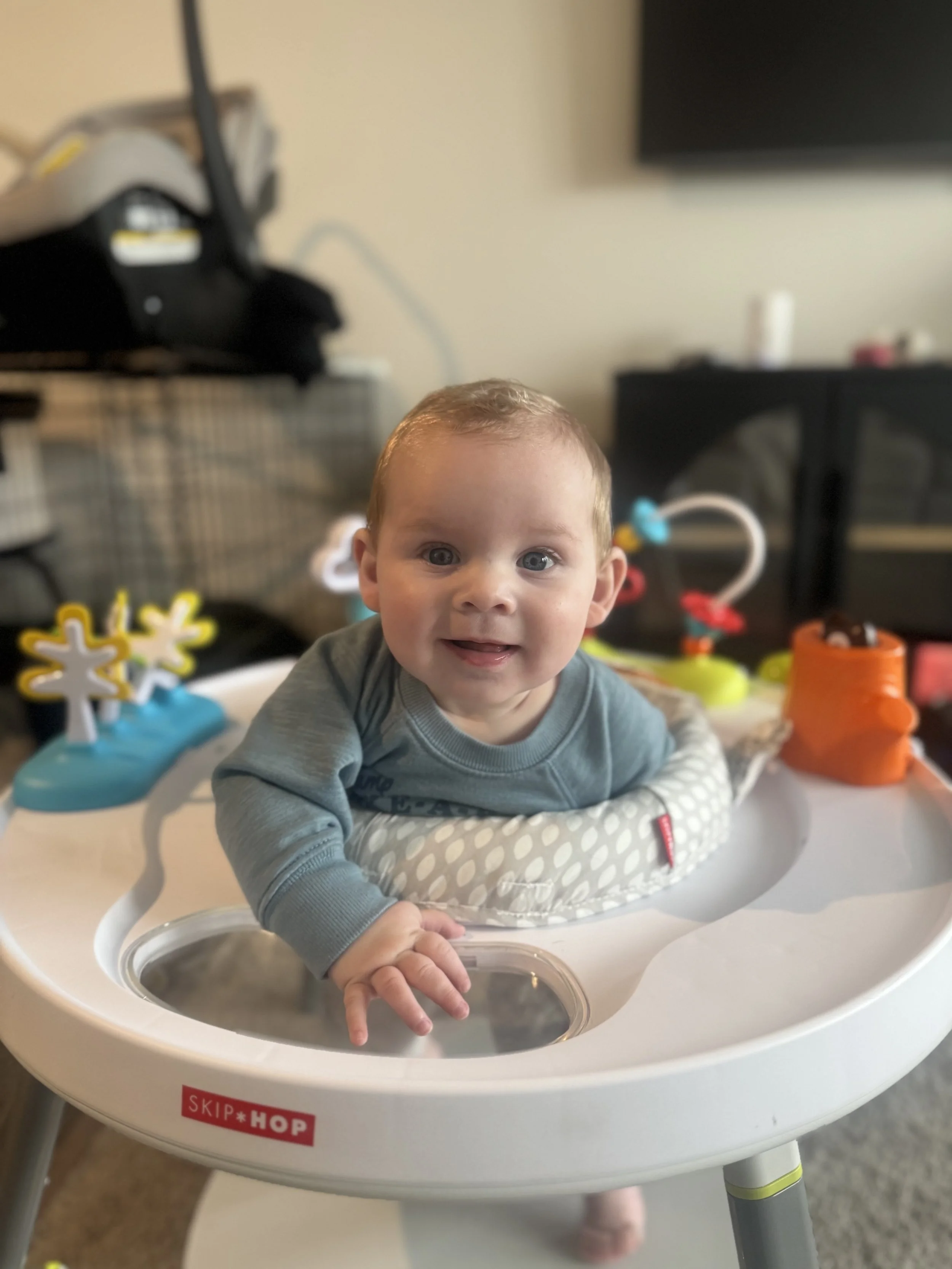 A baby with brown hair and blue eyes sitting in a white Skip Hop high chair, smiling and reaching out with one hand. The background shows a blurred living room with toys and furniture.
